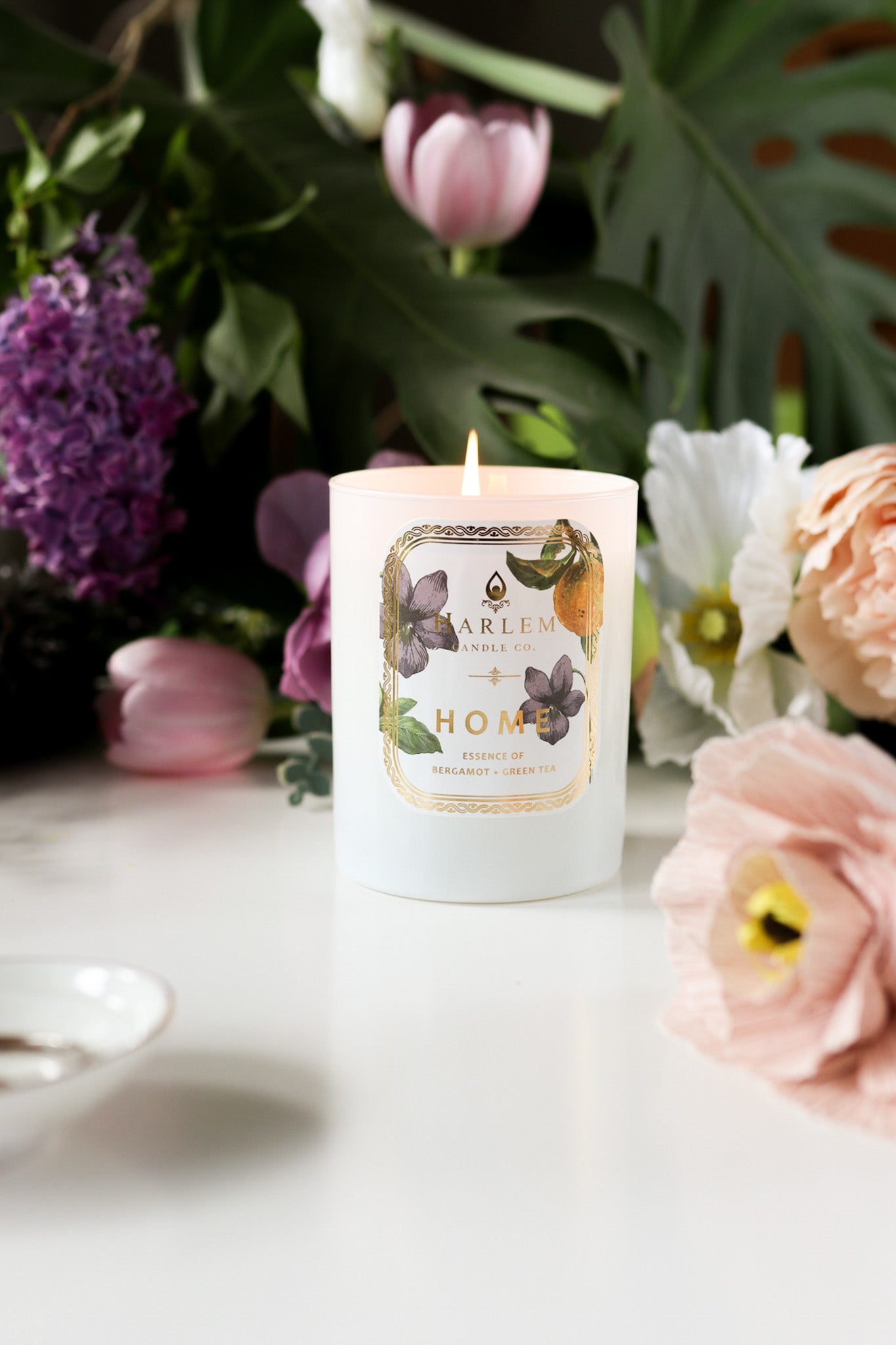 This is a vertical lifestyle image of the "Home" Luxury Candle with colorful flowers in the background.