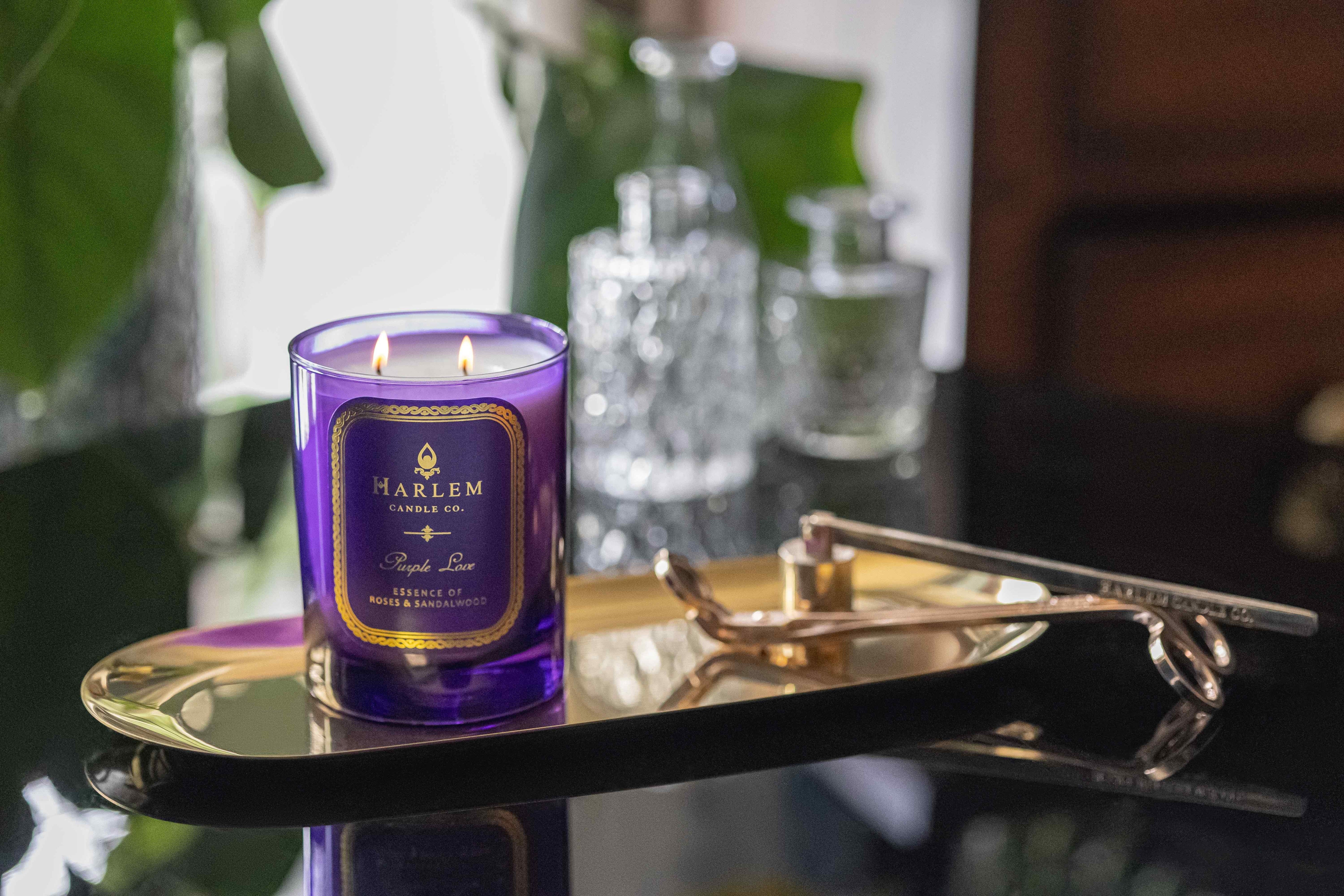 Lifestyle image of the purple love candle that is lit up. On a black table next to the Harlem Candle wick trimmer and snuffer with clear glass bottles in the background.