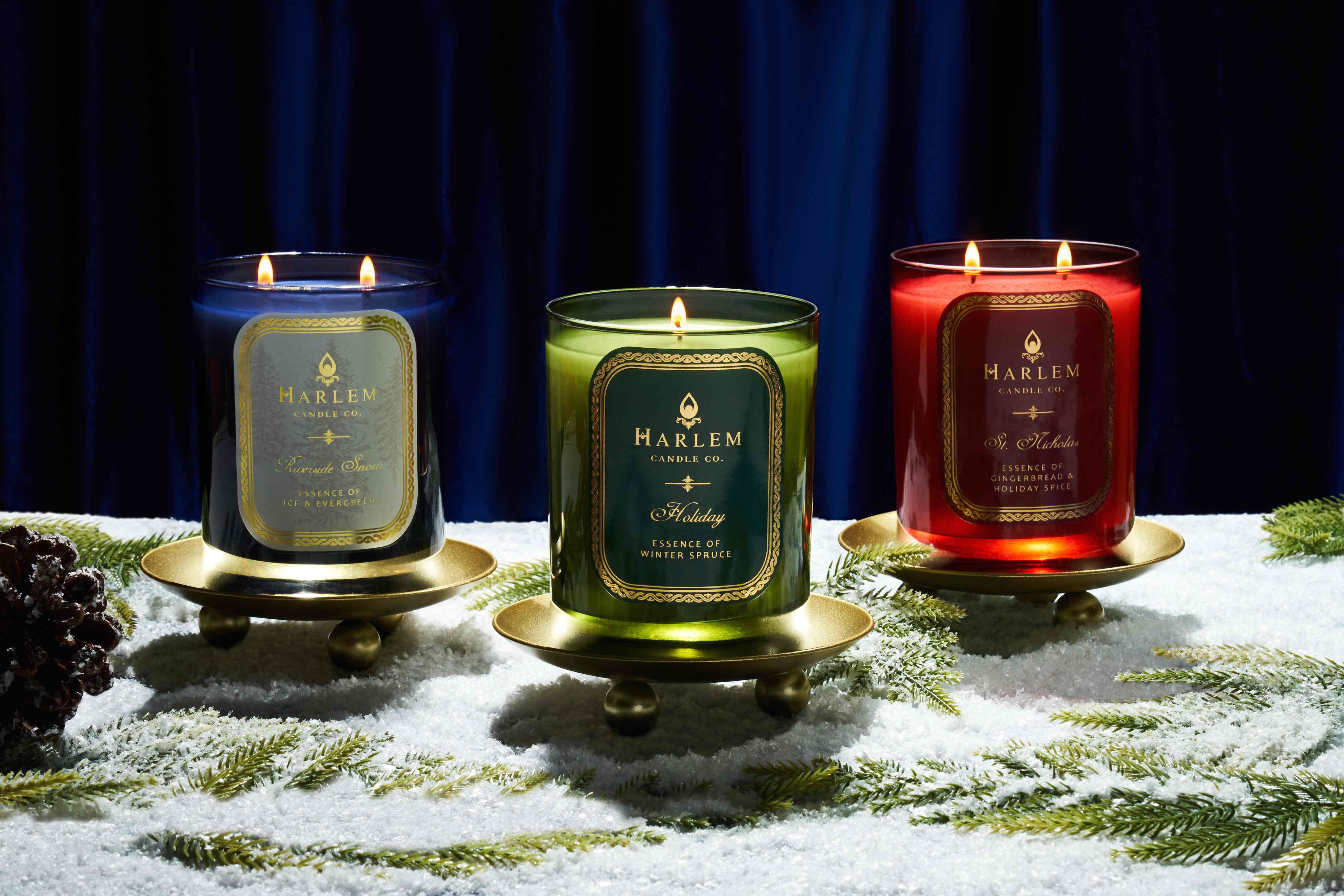 This is an image of our blue and white riverside snow to Wick candle, green Holiday, one wick candle, and our red Saint Nicolas two wick Candles.  These are pictured with a blue velvet curtain in the background with snow and pine leaves in the foreground.