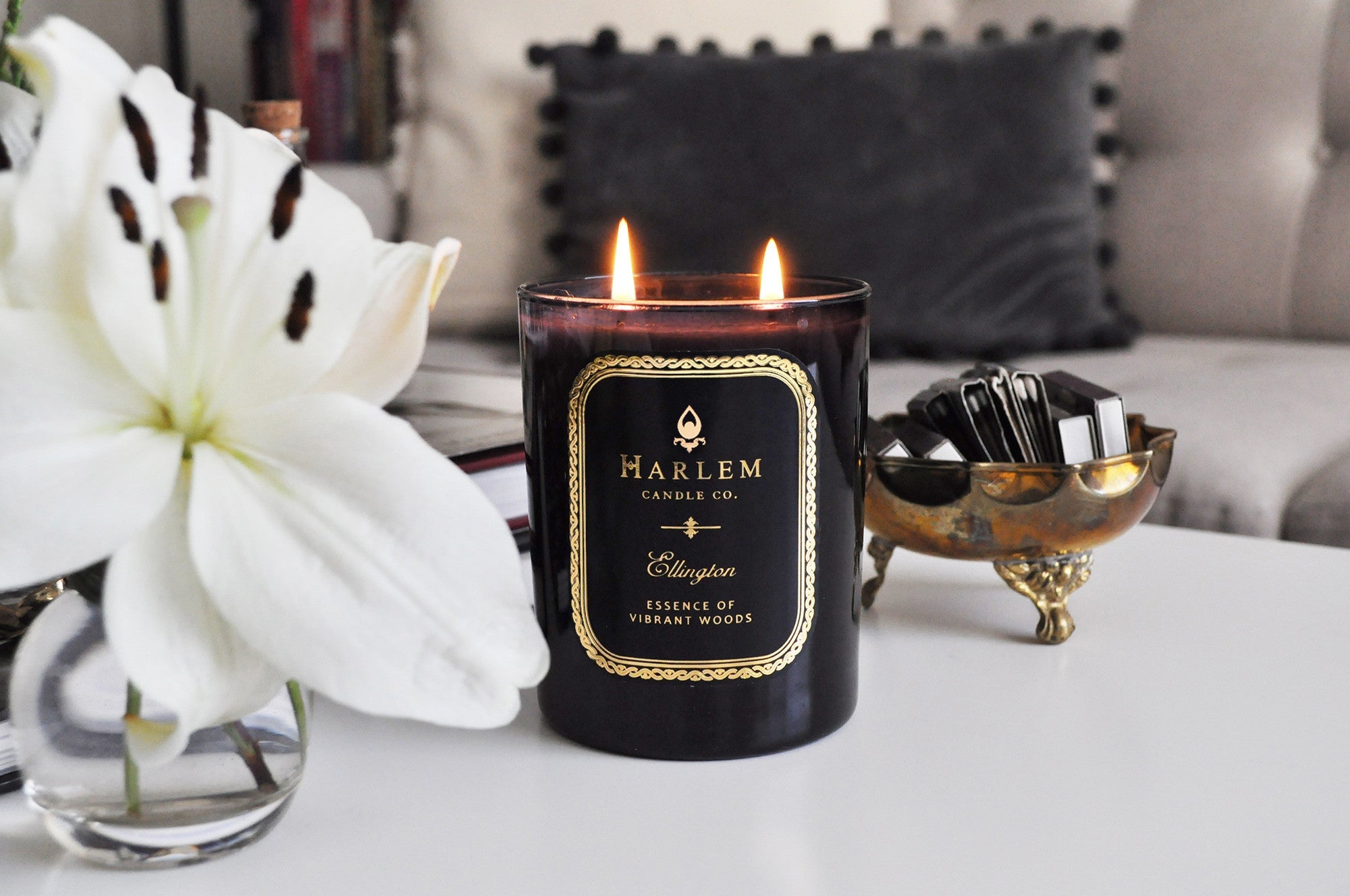 A stunning lifestyle image of our lit black Ellington 12 oz 2 wick candle sitting on a coffee table next to a vase of white flowers, a gold bowl of matches with a couch and black pillow in the background.