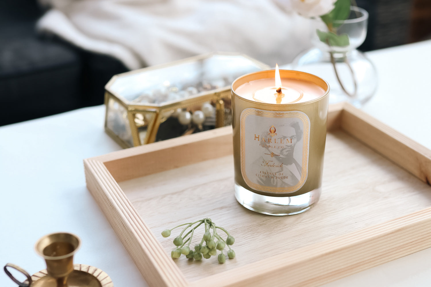 A beautiful lifestyle image of our lit 12 oz Frederick candle sitting on a wooden tray with gold jewelry and gold candle holders.