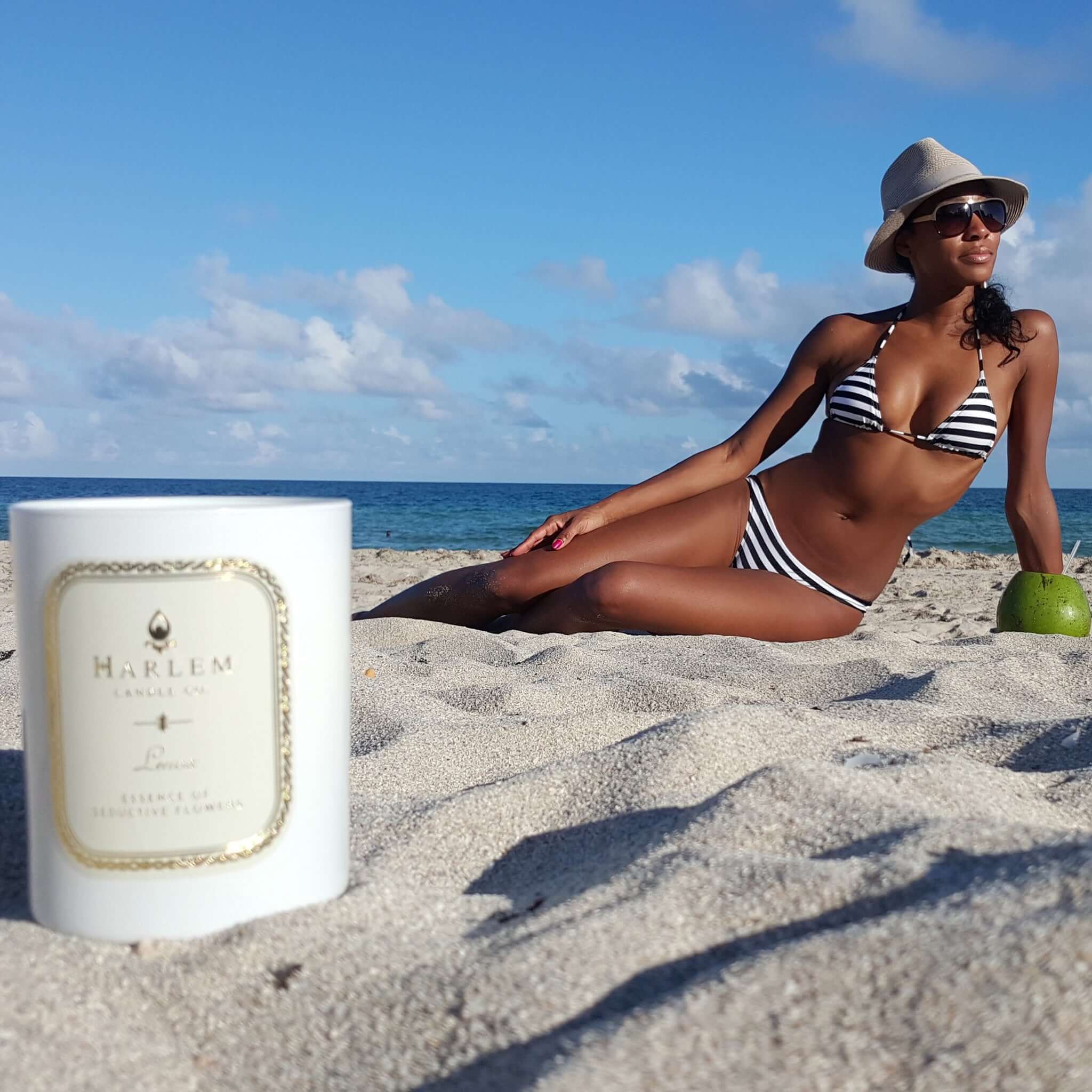 This photo is a lifestyle image of our Lenox 11 oz Candle in the sand at the beach with our stunning founder sunbathing.