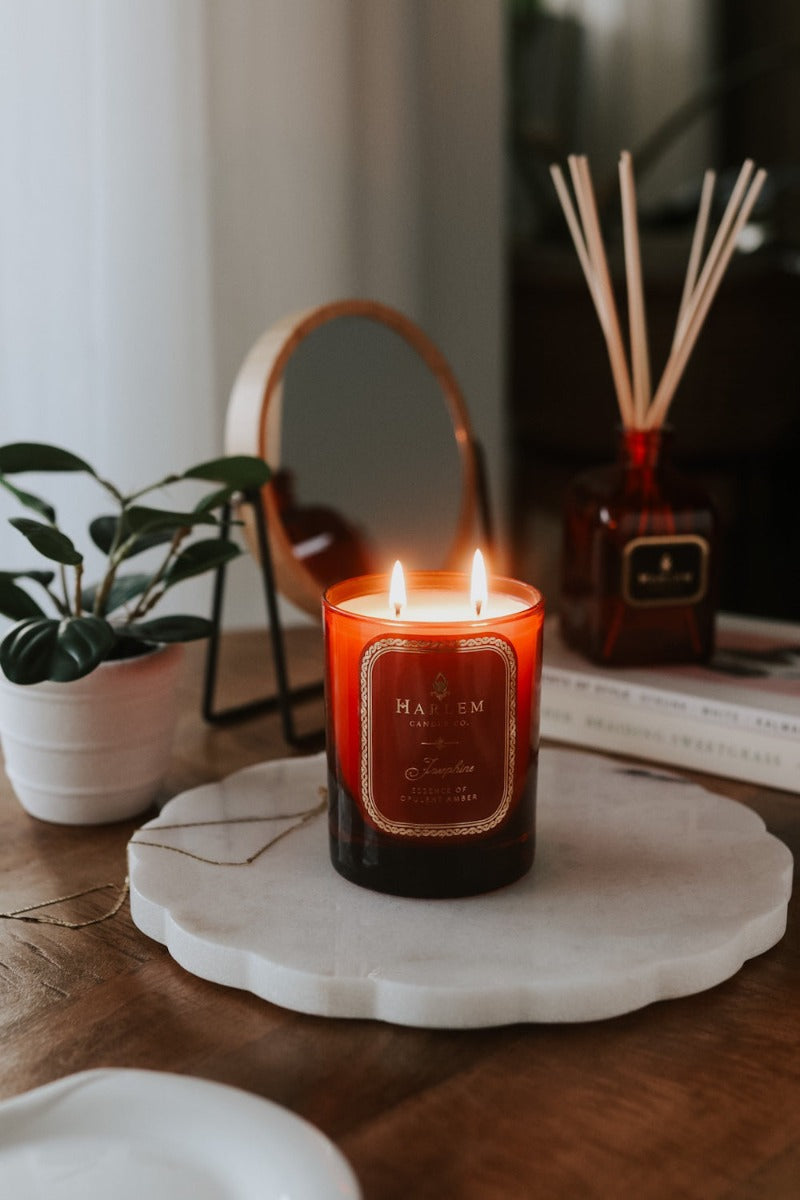 Our 11oz. 2-wick Josephine Candle in a red glass vessel sitting on a wooden table, next to a plant with the Josephine diffuser in the background.