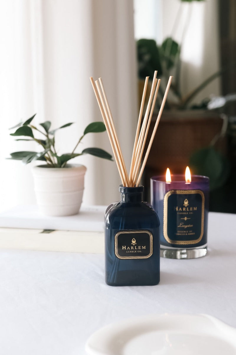 Our 8 fluid oz Langston Reed Diffuser with blue glass and reeds placed in diffuser glass sitting on a white table next to a 11oz Langston Candle