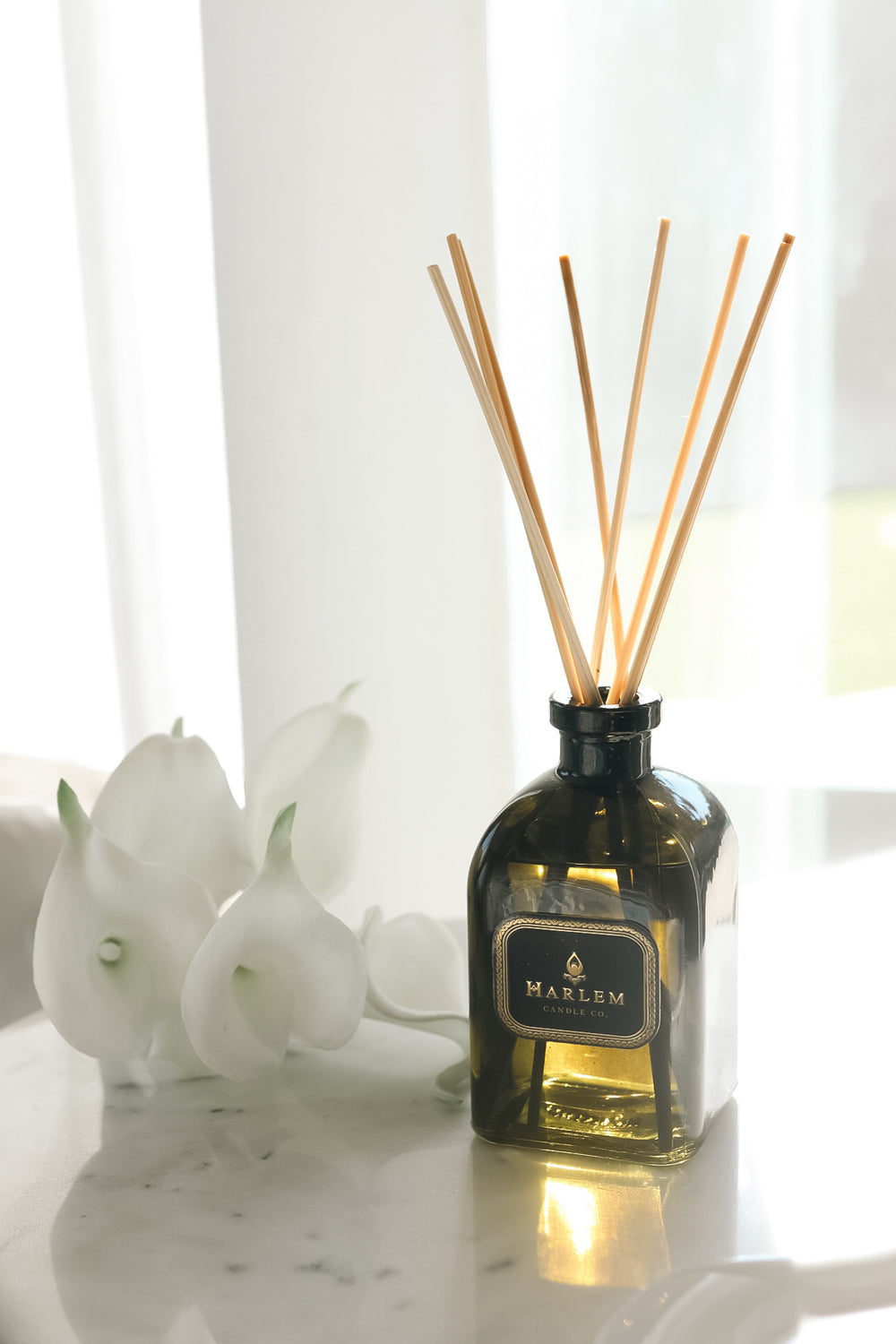 Our 8 fluid oz. Holiday Reed Diffuser with reeds, in a green glass vessel sitting on a white table next to flowers.