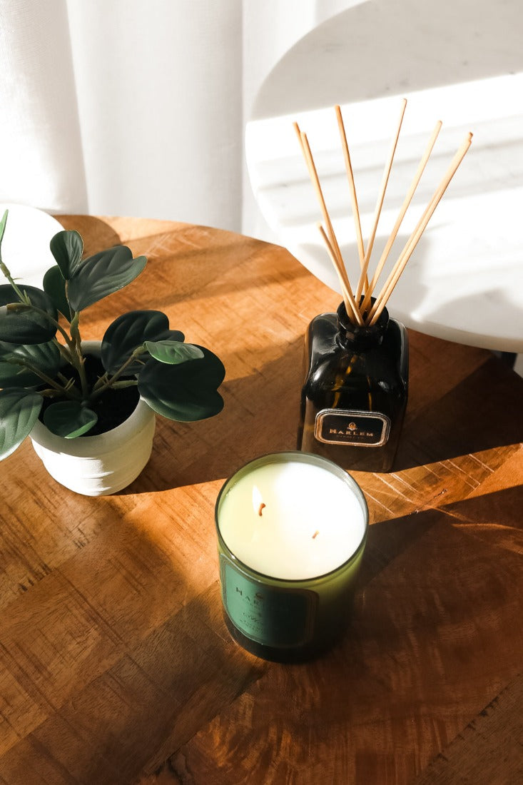 Our 8 fluid oz. Holiday Reed Diffuser with reeds, in a green glass vessel next to a green Holiday 11oz candle. sitting on a brown table..