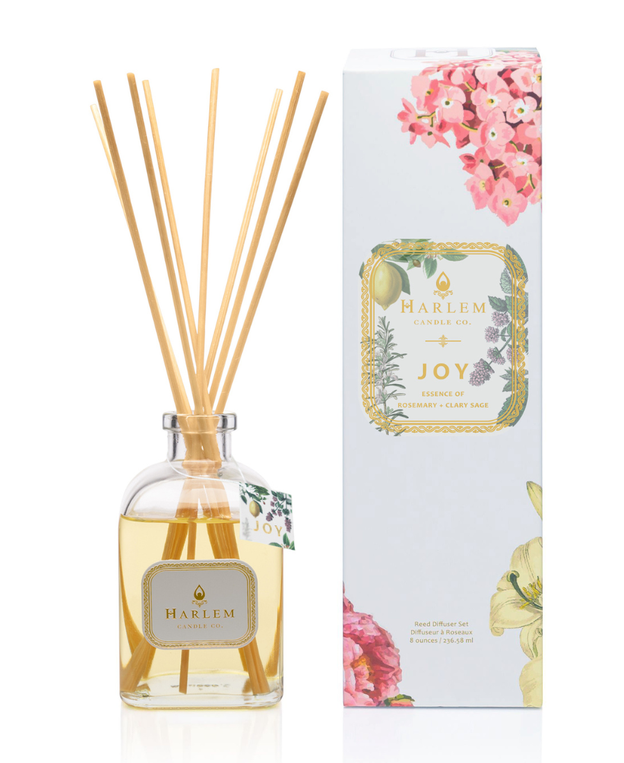This is an image of our Joy reed diffuser in a clear bottle with a white metallic label positioned next to the white floral decorative box.  The diffuser has a hang tag that says Joy to indicate that it is different from the other Botanical diffusers.