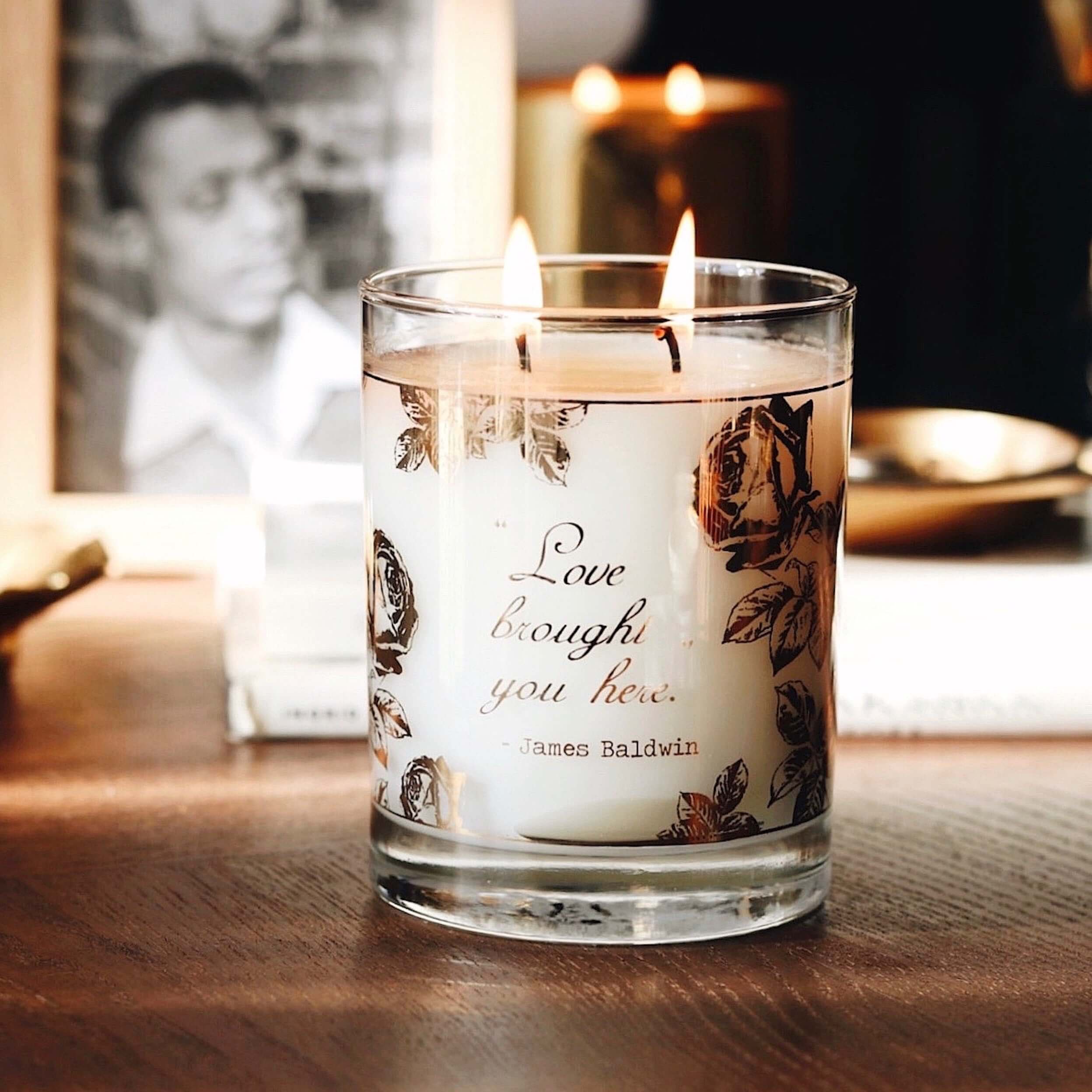 A lifestyle image of our 22k Gold James Baldwin "Love" candle, lit, on a wooden table in front of a black and white photo of James Baldwin in a gold frame.
