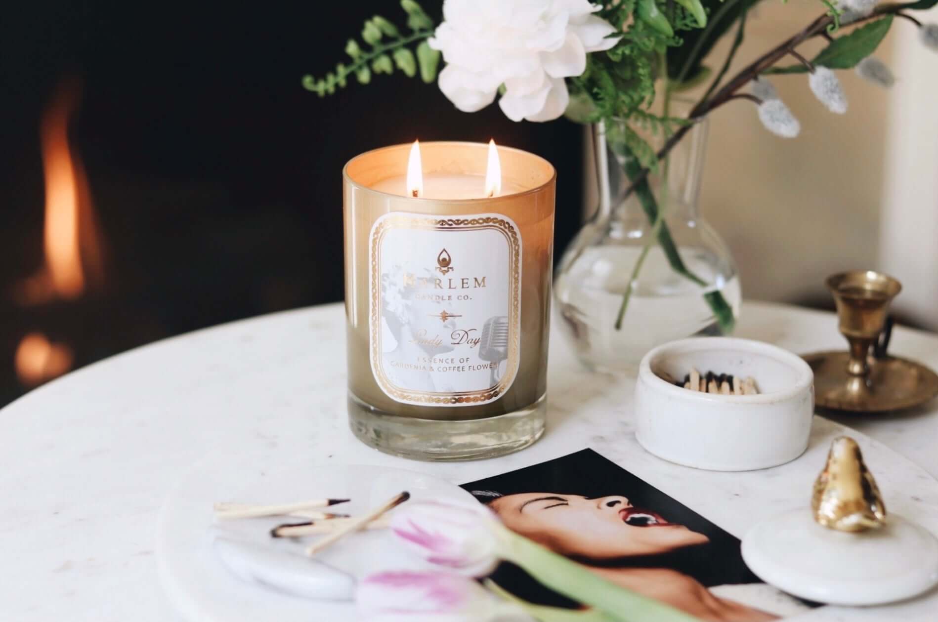 A lifestyle image of our Lady Day Gold 12 oz 2 wick, lit candle, on a white nightstand with photographs of Billie Holiday and greenery surrounding, and a gorgeous fire in the fireplace in the background.