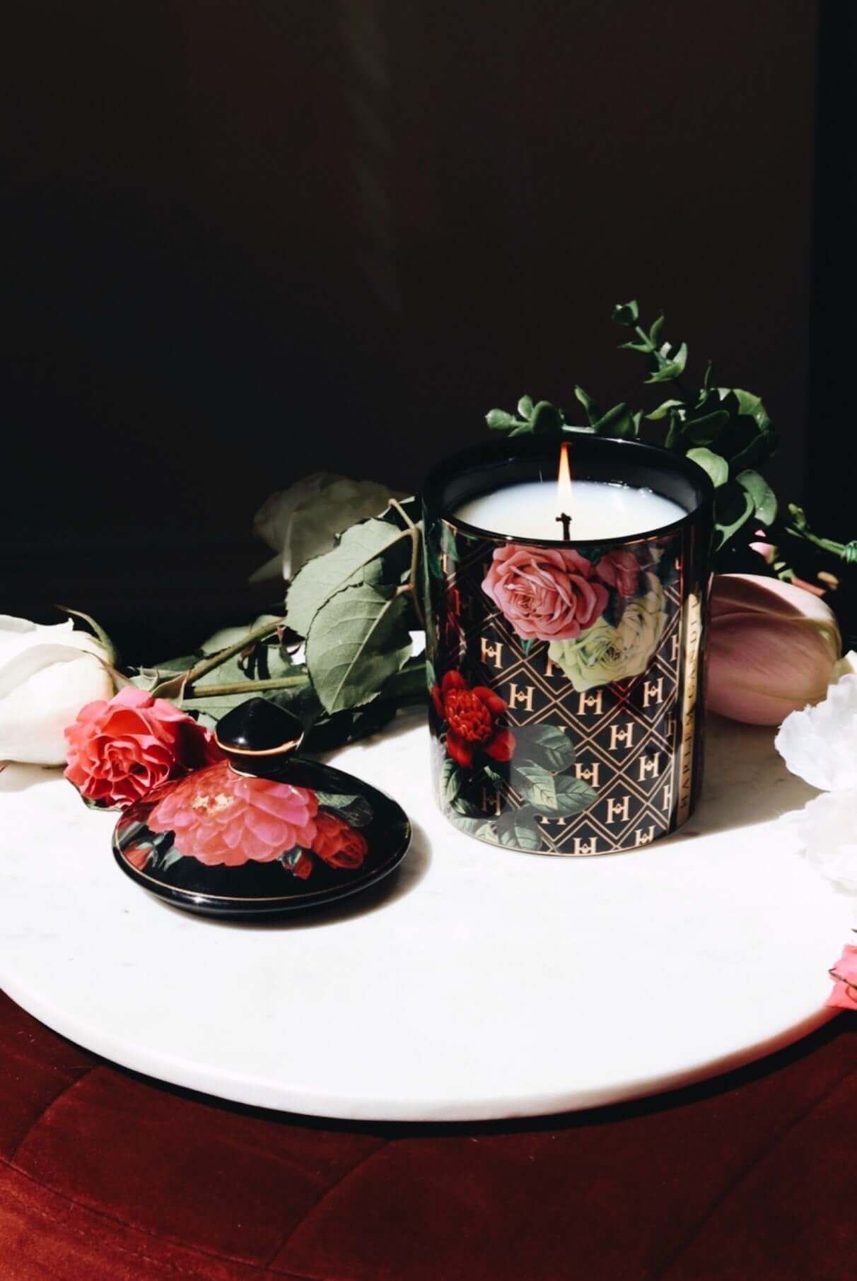 A lifestyle image of our "Love" Black Floral Ceramic Candle, the candle is lit on a white table with flowers and the lid next to the candle.