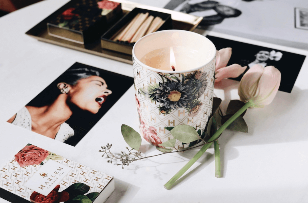 A lifestyle image of our Our “Lady Day” White Floral Ceramic Luxury Candle with lid, lit and on a white table next to matches and flowers and various images of Billie Holiday next to it.