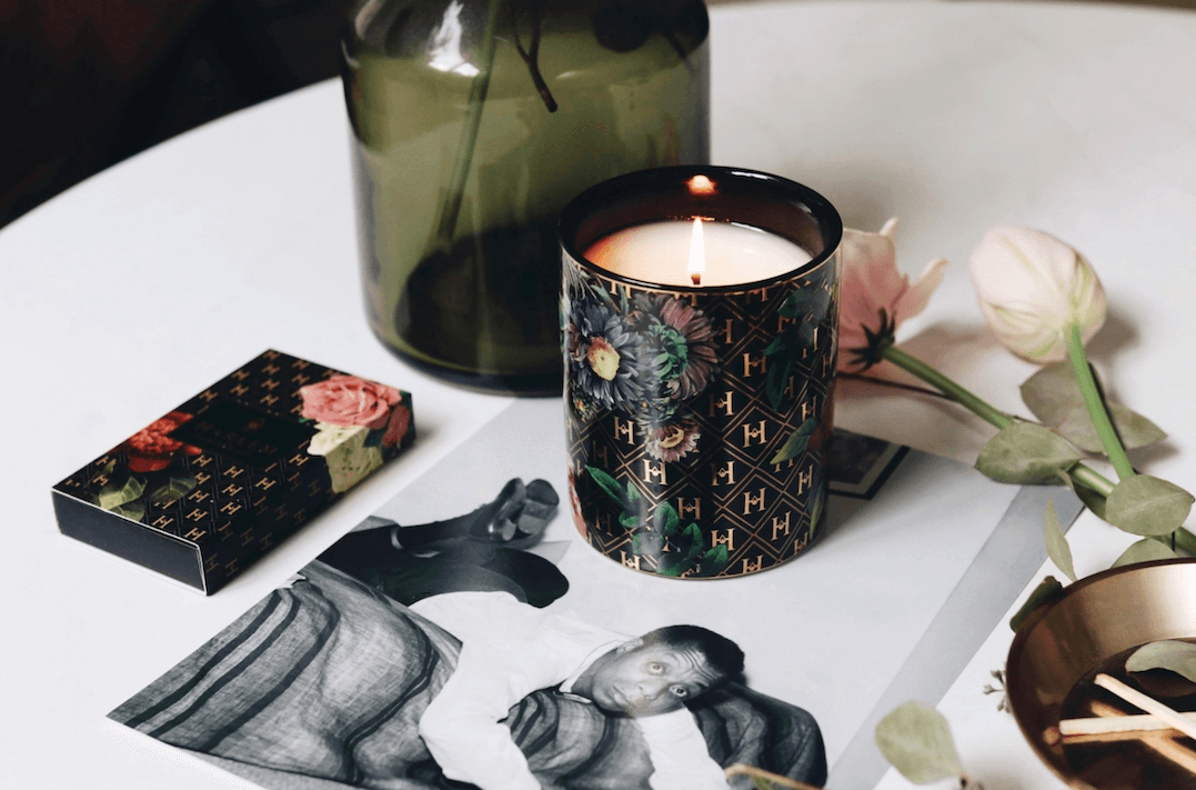 This lifestyle image features our Love Ceramic candle lit with matches to the side along with flowers, a black and white photograph of James Baldwin on a white table.