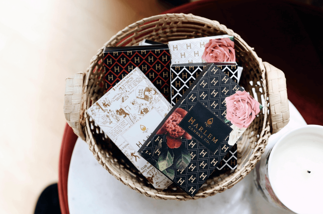 An array of our match boxes in black, red gold and white arranged in a basket sitting on a white side table next to a candle.