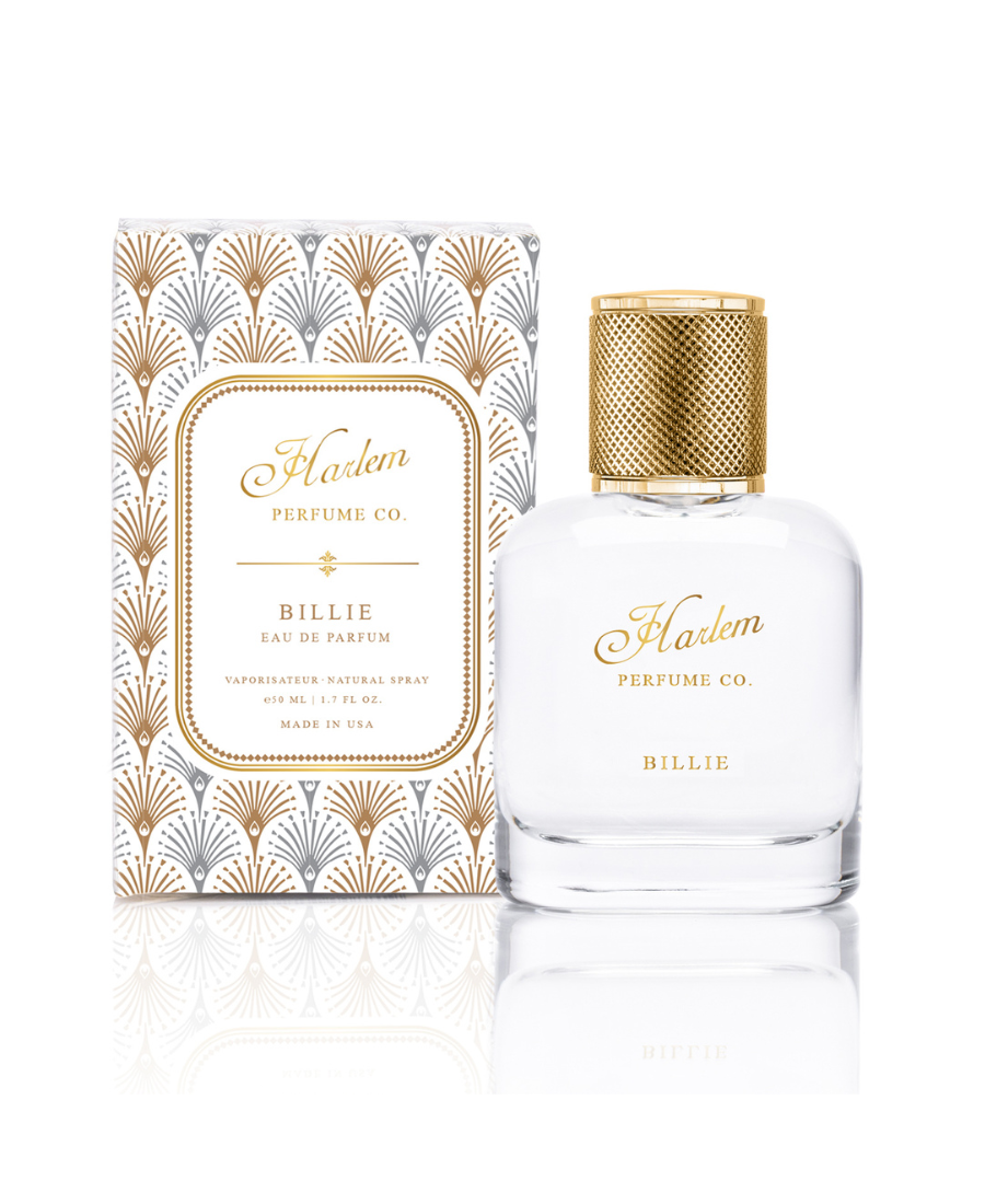 This is an image of our Billie eau de Parfum bottle against a white background. The bottle is clear with gold letters that say Harlem Perfume Co and the word Billie as the name of the fragrance. This bottle is pictured next to its decorative box.