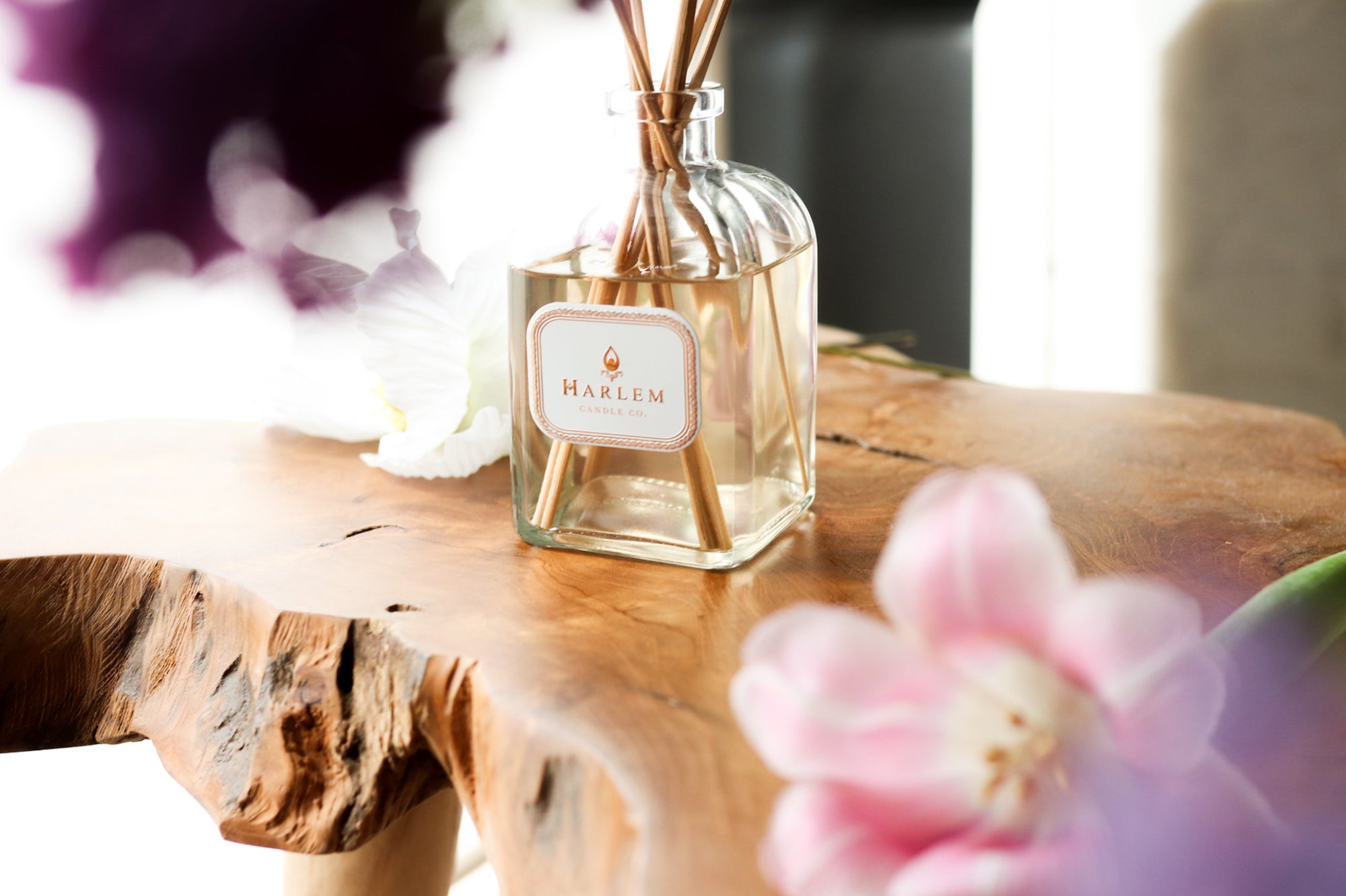 This is an image of our dream reed diffuser in a life style setting with the flowers surrounding it. It is on a wooden surface with a white and gold metallic label on the clear bottle.