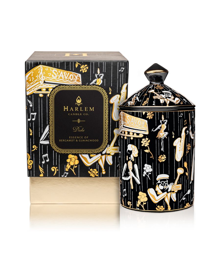 This is an image of our black gold and white Duke, ceramic candle with a lid. It features illustrations of Billie Holiday, the savoy ballroom, Duke Ellington, and other original illustrations.  This candle is featured next to its decorative box.