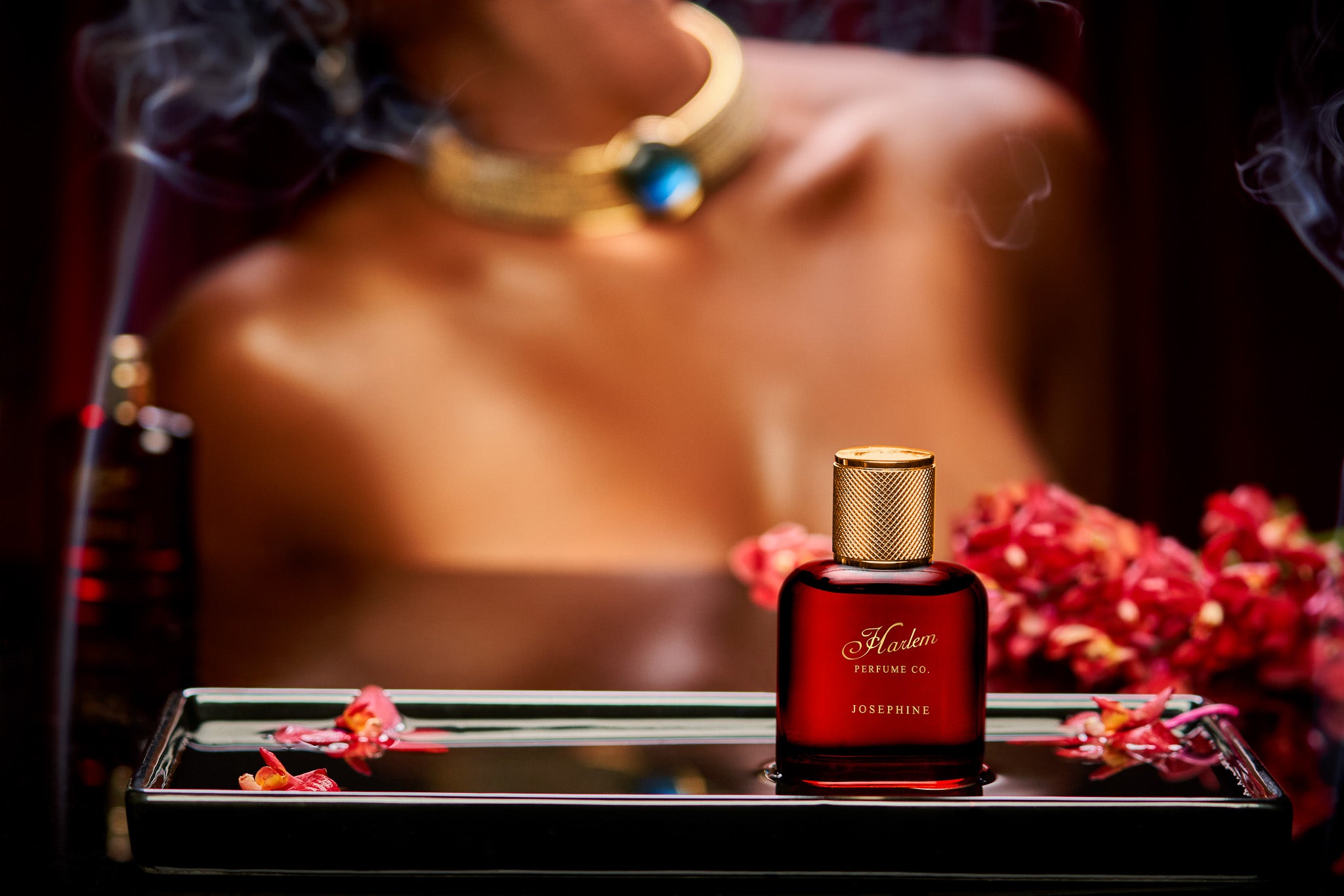This is a lifestyle image of our red Josephine Perfume bottle with a woman in the background, wearing a gold necklace with incense blowing around her.