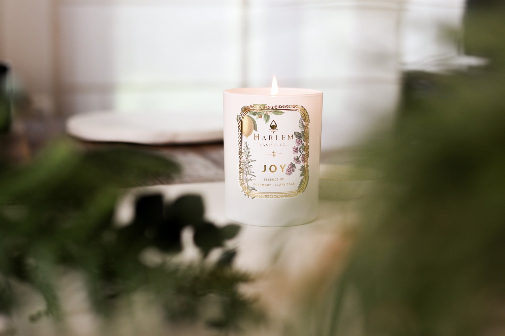 This is an image of the "Joy" Luxury Candle on a wooden table with a white place mat and greenery in the foreground.