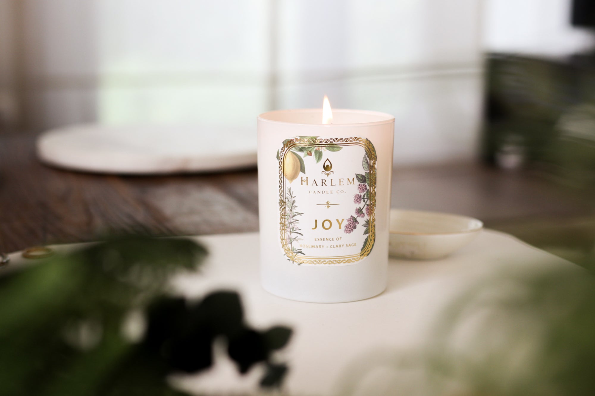 This is an image of the "Joy" Luxury Candle on a wooden table with a white place mat and greenery in the foreground.