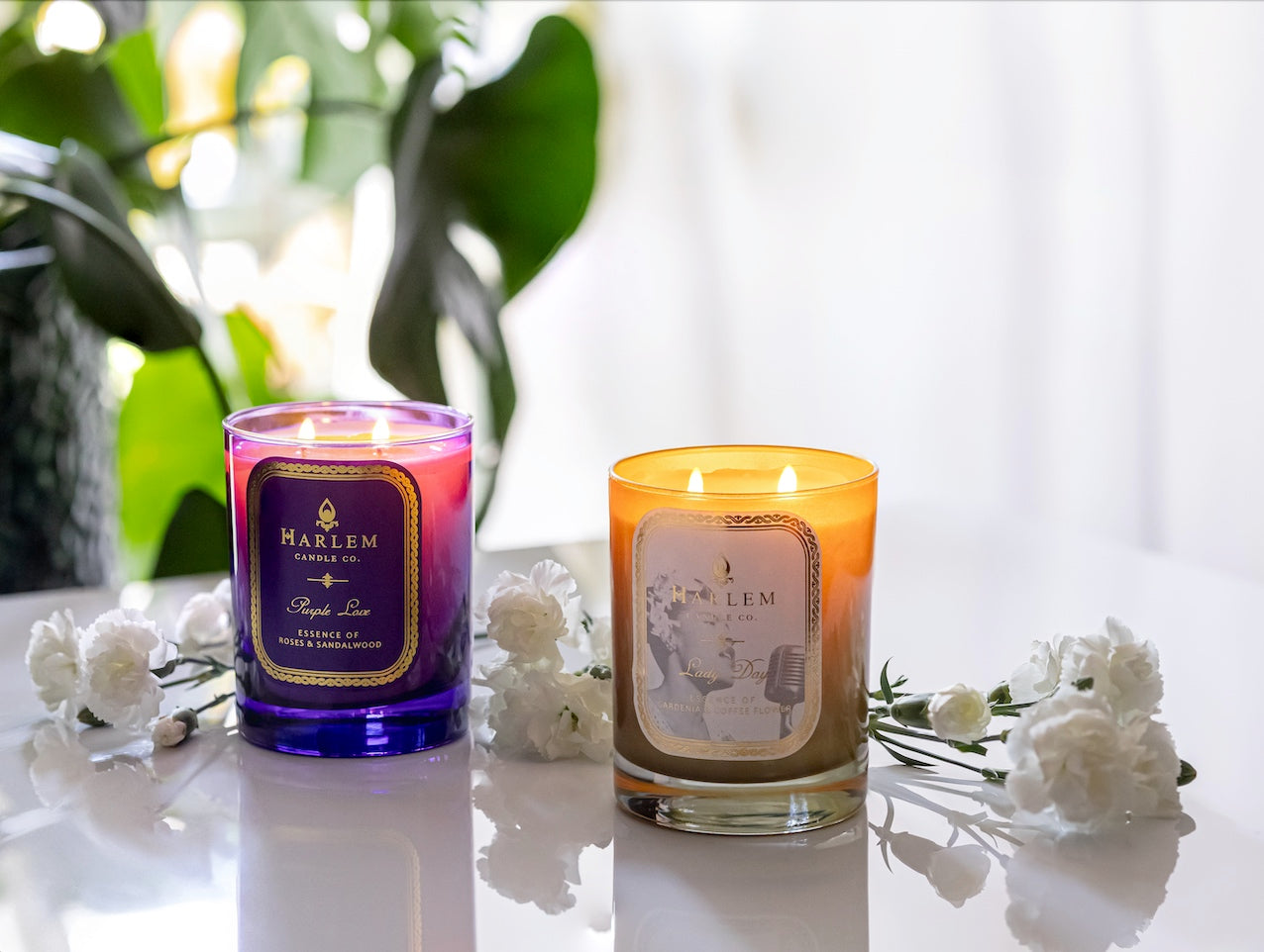  This is an image of our Purple Love and gold and white Lady Day candles in a lifestyle setting where they are surrounded by green plants and white flowers.