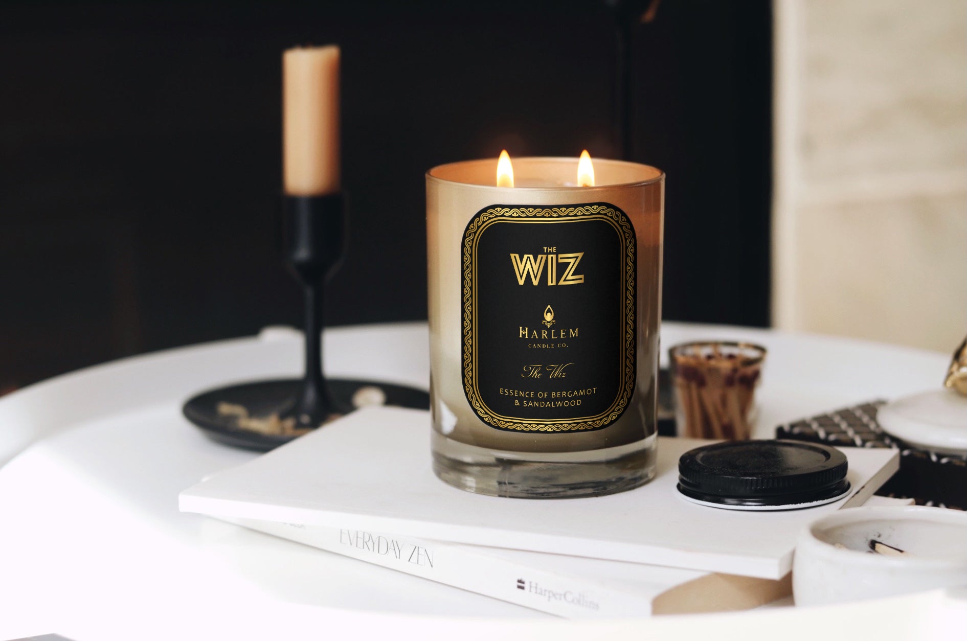 This is an image of the wiz candle in a lifestyle setting. It is pictured next to a gold and black taper candle with matches and books surrounding it.