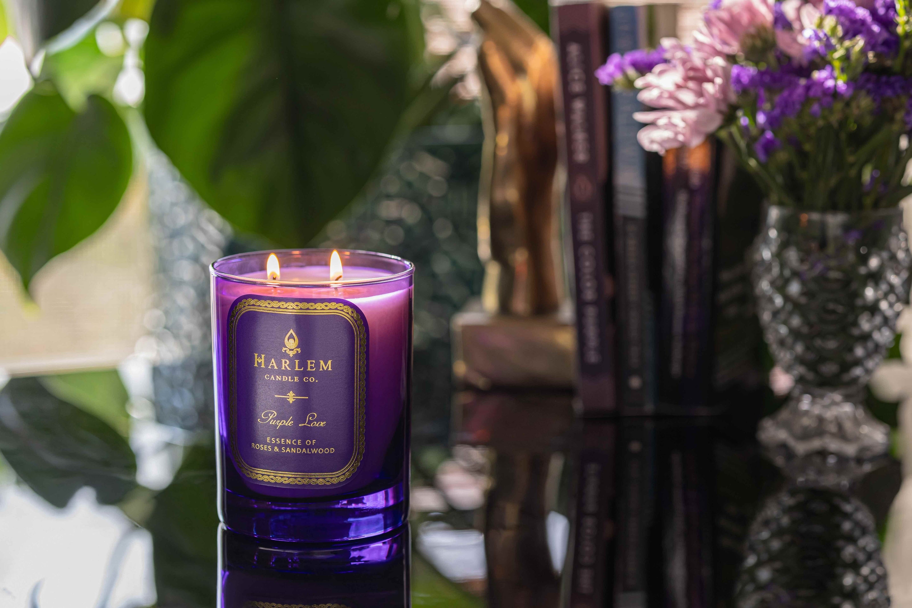 This is a lifestyle image of our purple love candle with green leaves, plants, books, and purple flowers in the background