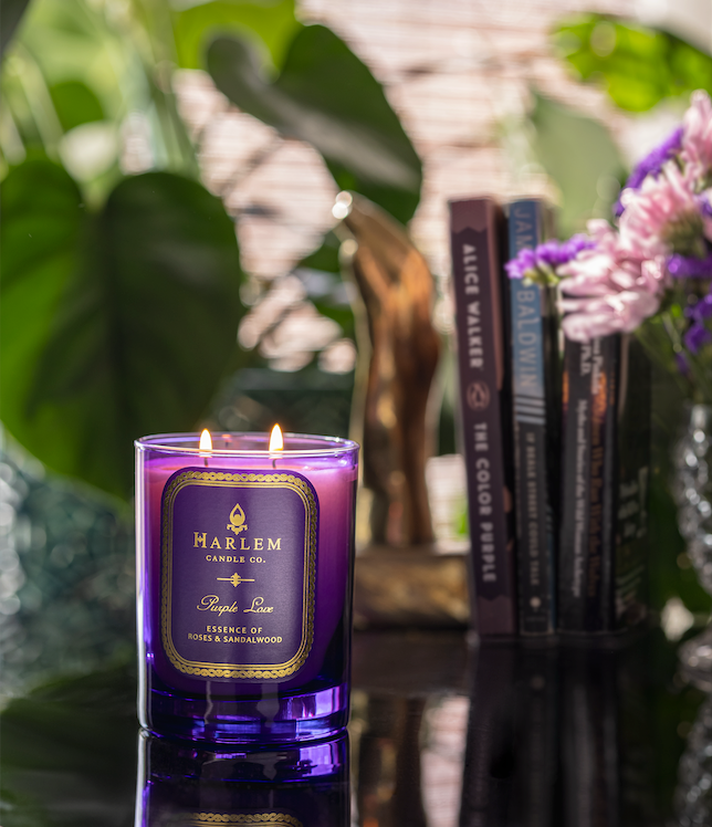Lifestyle image of the purple love candle that is lit up. On a black table with purple flowers and books in the background.
