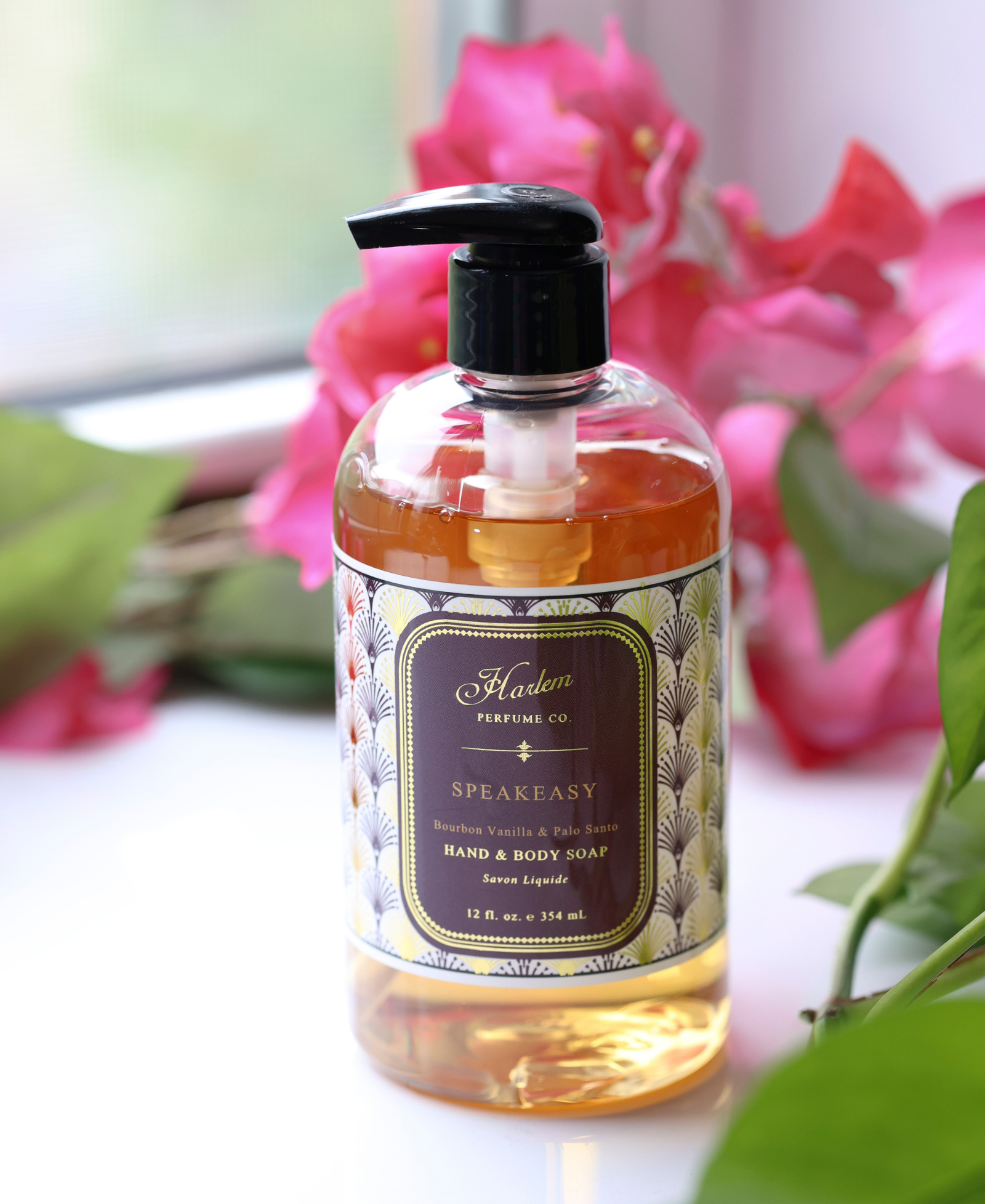 This is an image of our Speakeasy Soap pictured next to pink flowers.  The bottle has a black pump and the label has a unique Art Deco print with the details of the soap/
