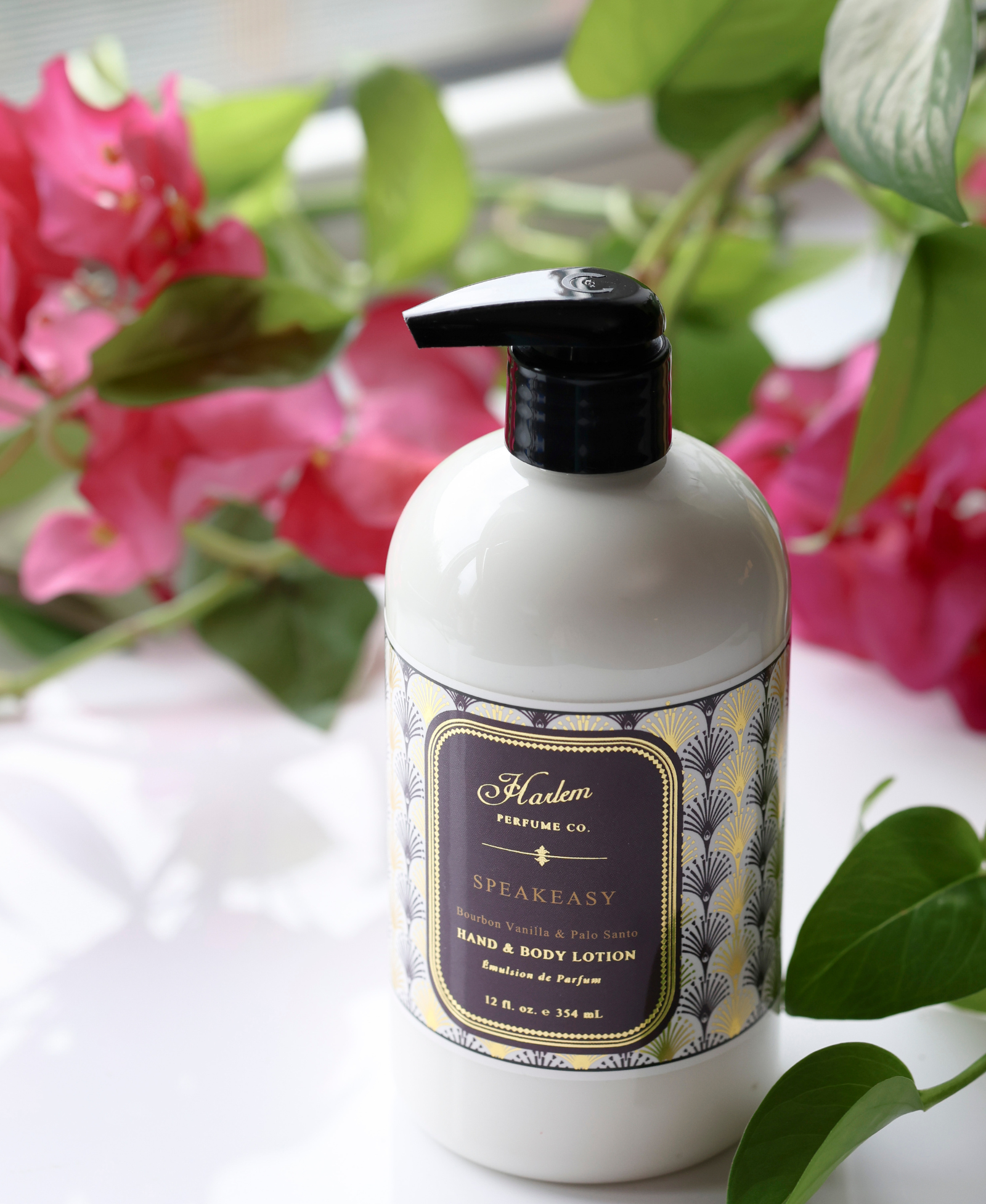 This is an image of our Speakeasy lotion pictured next to pink flowers.  The bottle has a black pump and the label has a unique Art Deco print with the details of the lotion