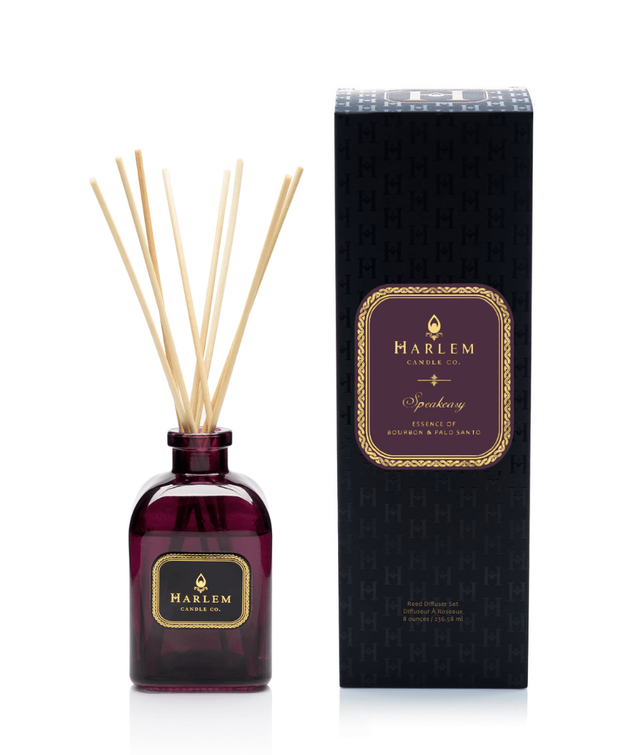 Our 8 fluid oz. Speakeasy Reed Diffuser with reeds, in a purple glass vessel sitting next to its decorative box on a white background.