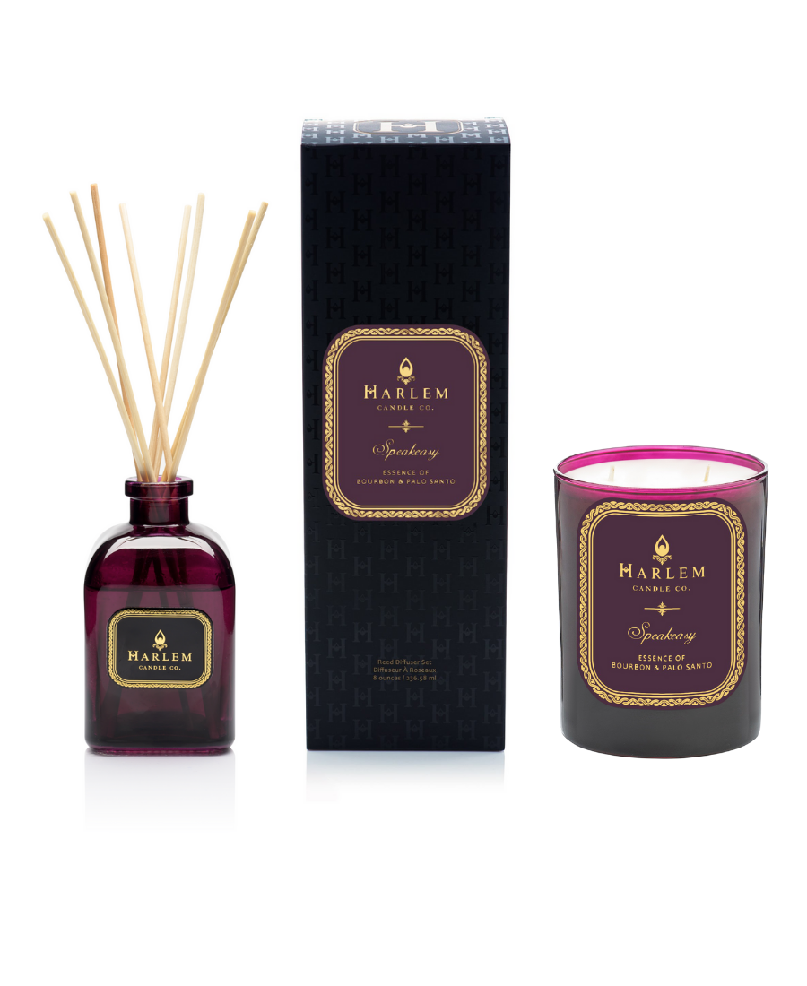 Our 8 fluid oz. Speakeasy Reed Diffuser with reeds,and 11oz Speakeasy Candlein a purple glass vessel sitting next to its decorative box on a white background.