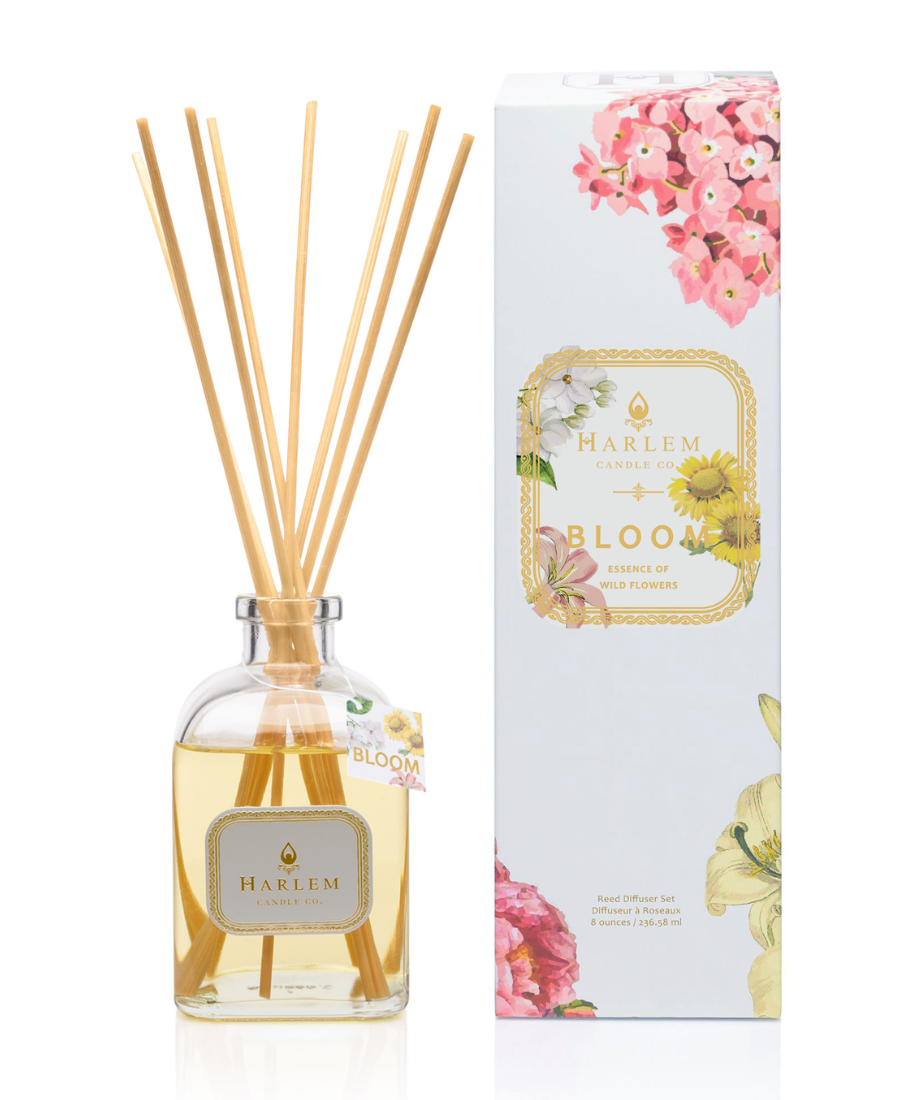 This is an image of our Bloom reed diffuser in a clear bottle with a white metallic label positioned next to the white floral decorative box.  The diffuser has a hang tag that says Bloom to indicate that it is different from the other Botanical diffusers.