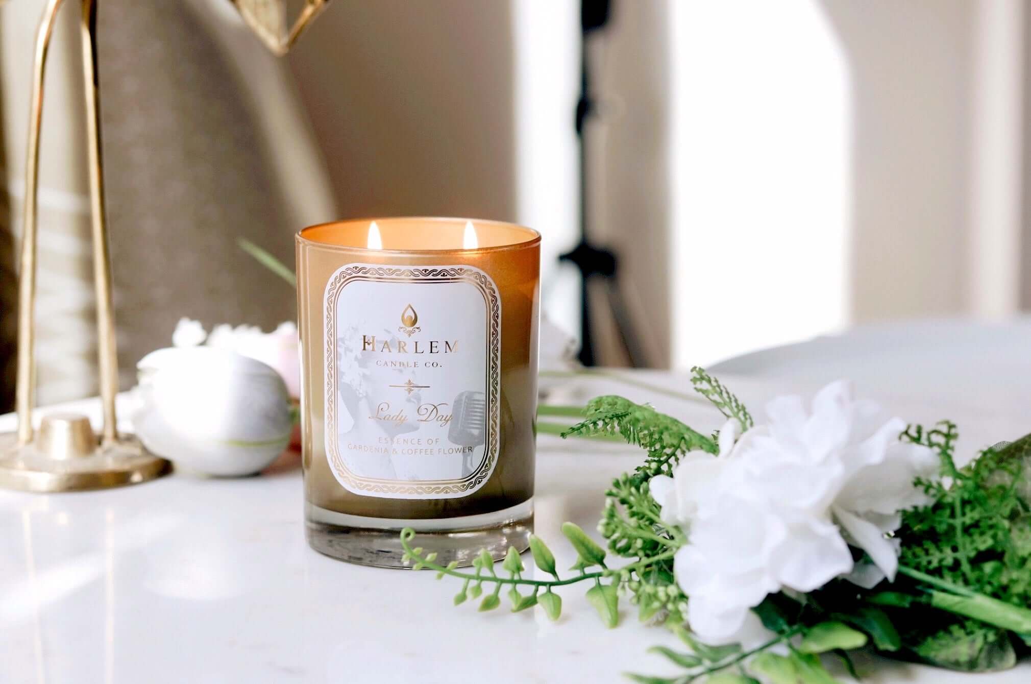 A lifestyle image of our Lady Day Gold 12 oz 2 wick candle, lit, sitting on a white table with greenery and flowers surrounding the gold candle.