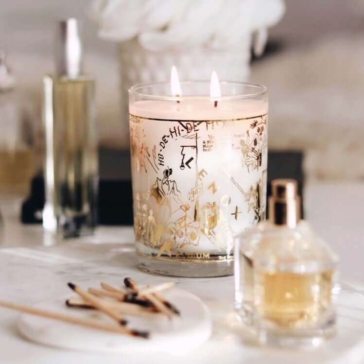 A gorgeous lifestyle image of our 22K Gold Nightclub Map Of Harlem Savoy Luxury Candle sitting on a white marble table with matches and glass perfume bottles surrounding the lit candle.