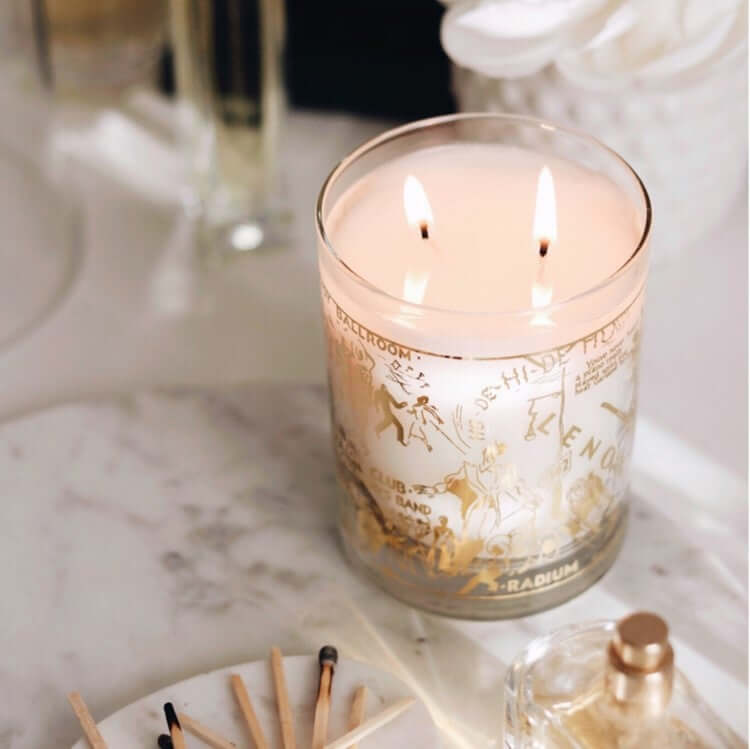 A gorgeous lifestyle image of our 22K Gold Nightclub Map Of Harlem Savoy Luxury Candle sitting on a marble slab next to matches and an elegant glass perfume bottle.
