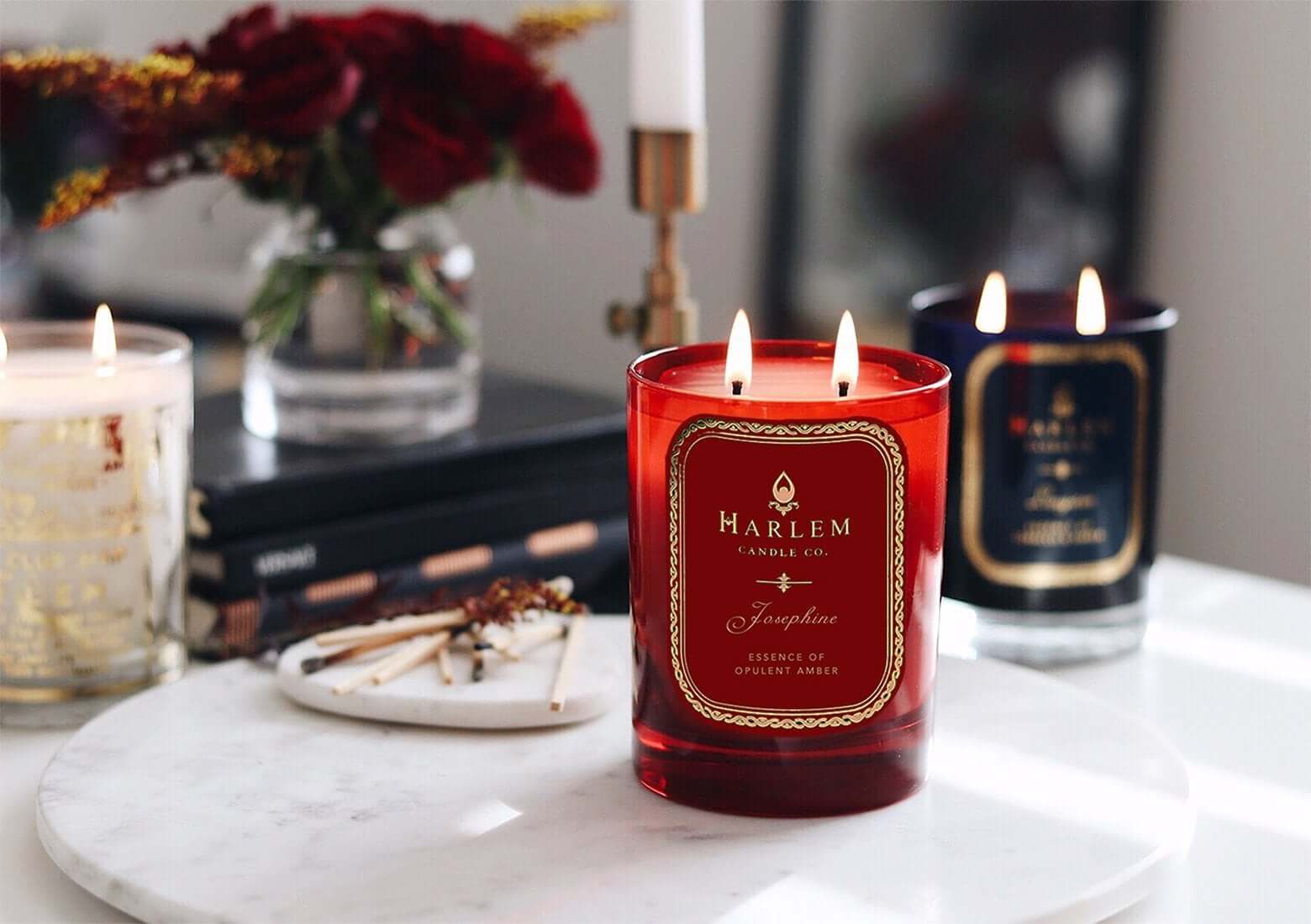 A lifestyle image with three lit candles.  The red Josephine 11 oz, the Langston 11 oz and a 22K Gold Cocktail glass on a marble table with flowers and gold candle holders in the background. 