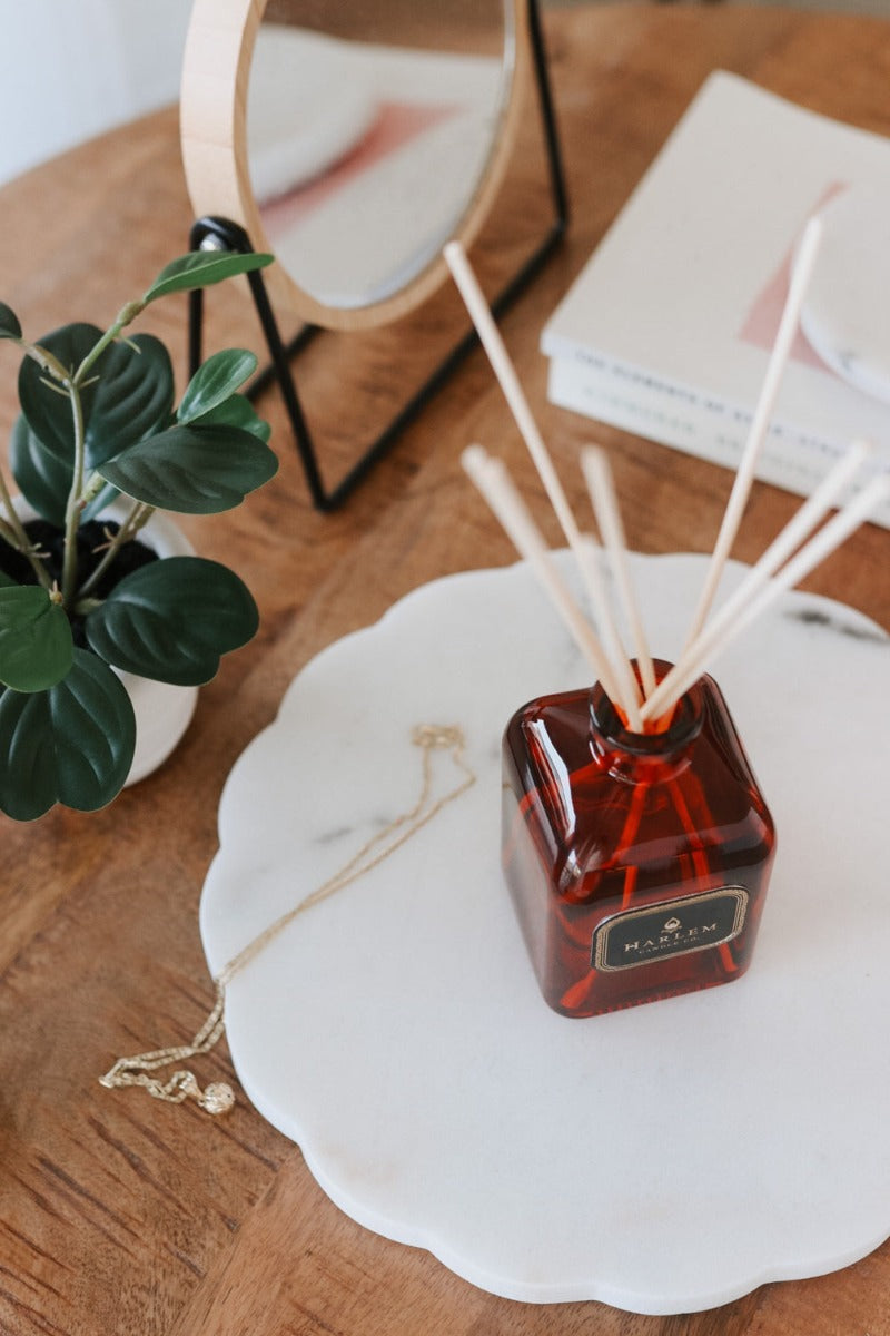 Our 8 fluid oz. Josephine Reed Diffuser with reeds, in a red glass vessel sitting on a wooden table, next to a plant and necklace.