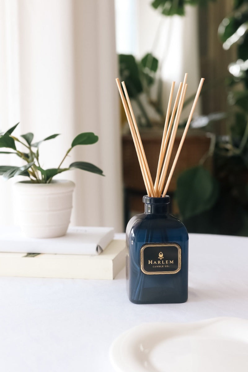 Our 8 fluid oz Langston Reed Diffuser with blue glass and reeds placed in diffuser glass sitting on a white table next to a plant..