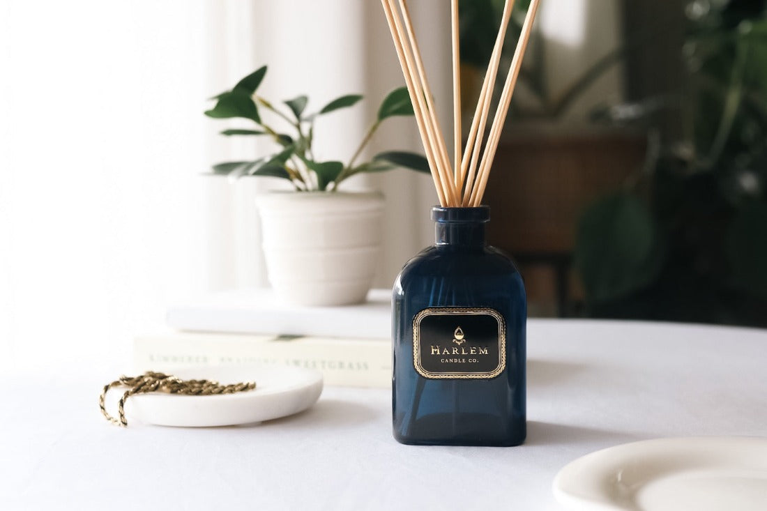 Our 8 fluid oz Langston Reed Diffuser with blue glass and reeds placed in diffuser glass sitting on a white table next to a plant.