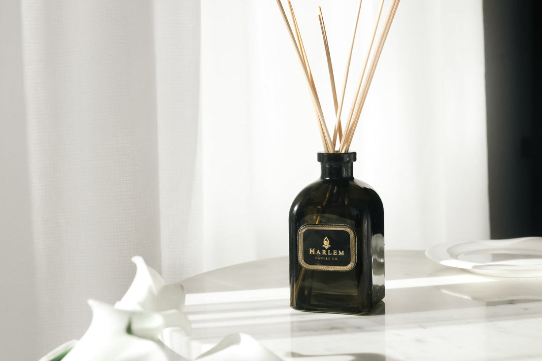 Our 8 fluid oz. Holiday Reed Diffuser with reeds, in a green glass vessel sitting on a white table next to flowers.
