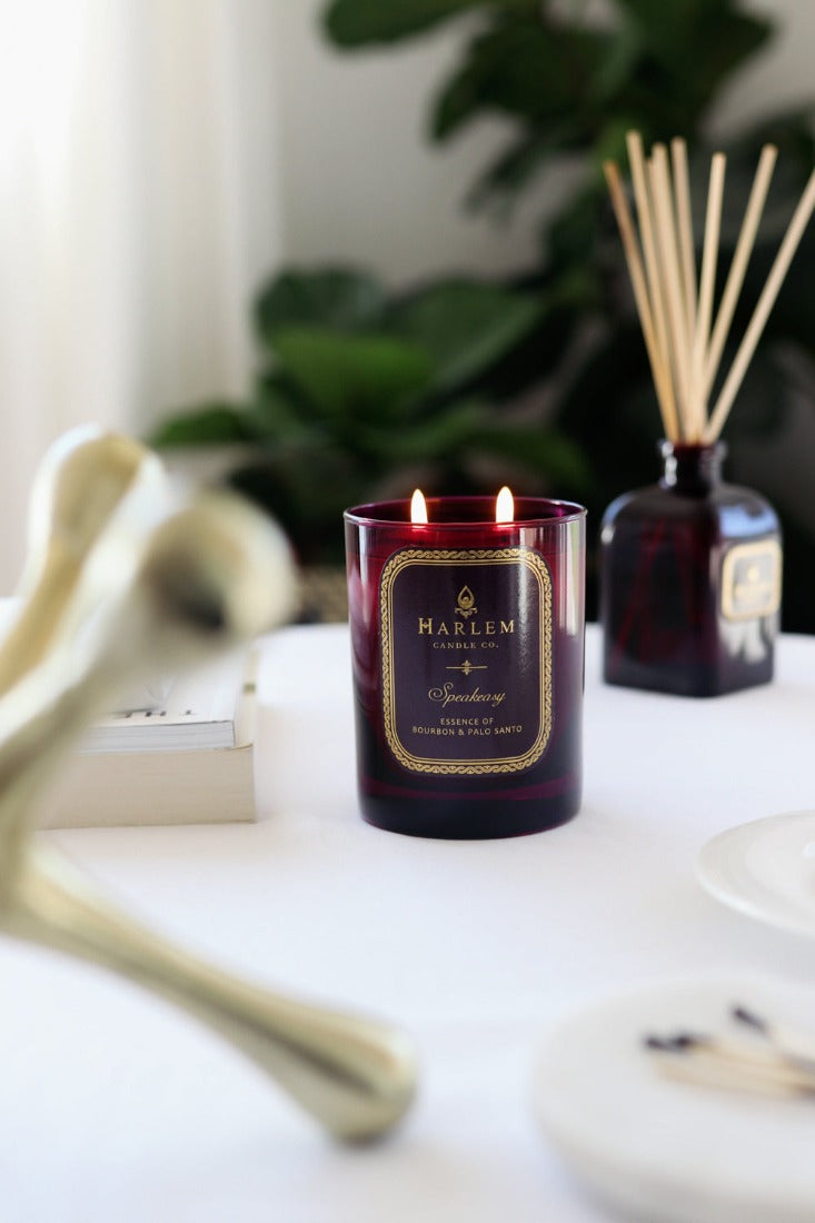 Our 8 fluid oz. Speakeasy Reed Diffuser with reeds,and 11oz Speakeasy Candlein a purple glass vessel sitting next to its decorative box on a white table.