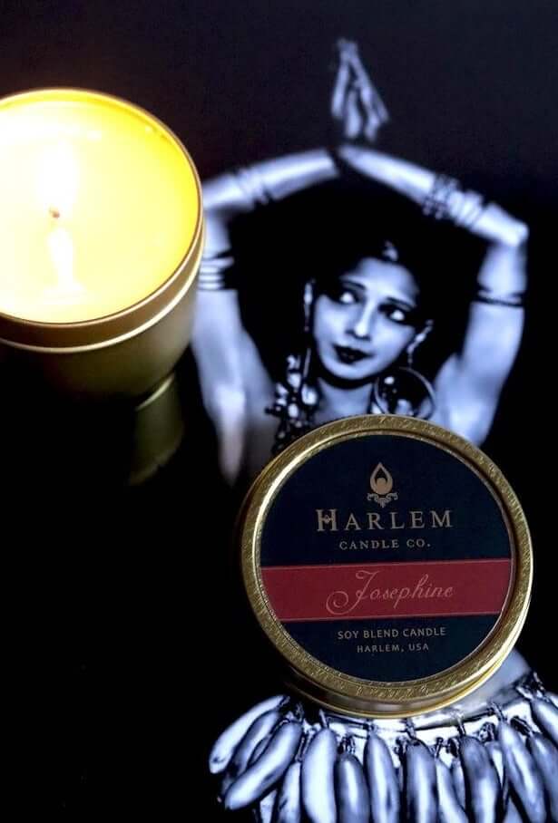 Our Josephine 4 oz travel candle in tin with top next to the soft glowing candle on a black background with a black and white image of Josephine Baker.