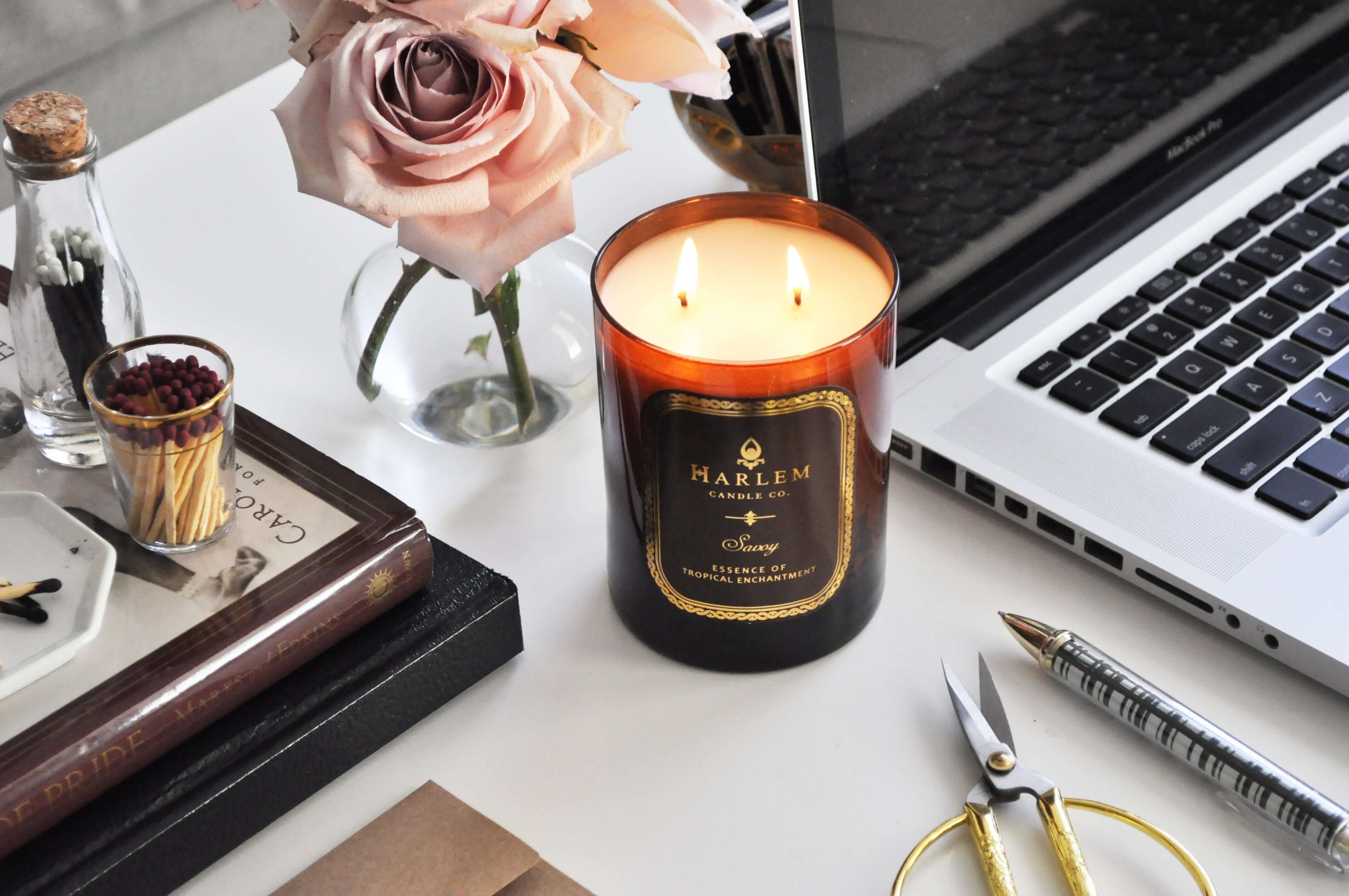 Our 12 oz Amber Savoy 2 wick, lit candle in a lifestyle image sitting on a desk next to a Mac Book Pro Laptop, some books, matches and some pink roses in a vase.
