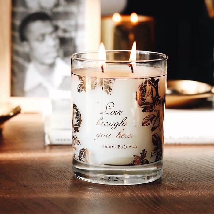 This is a lifestyle image of our Love Brought You Here James Baldwin lit 12 oz 2 wick candle on a wooden side table with a black and white photograph of James Baldwin in the background.