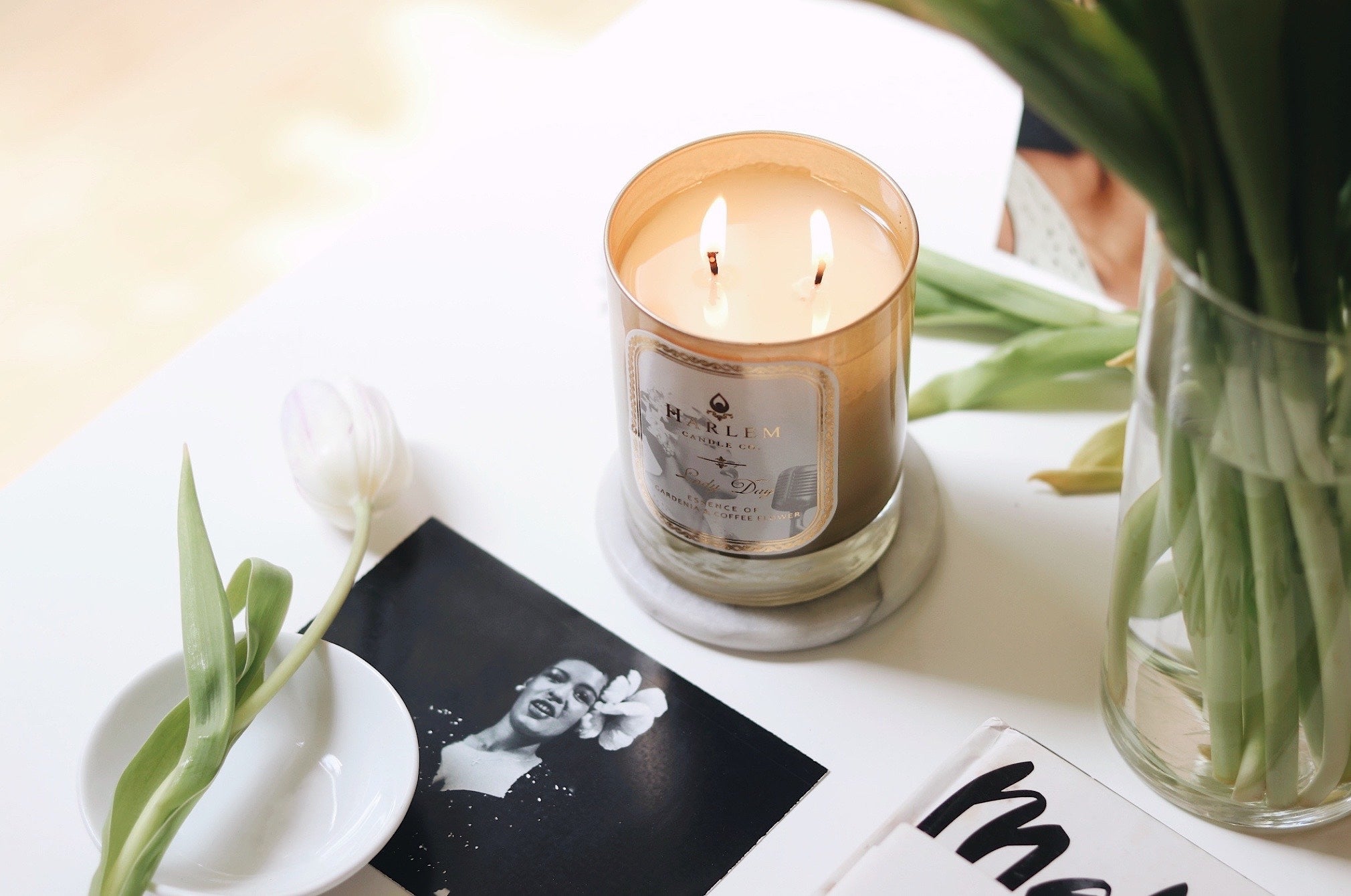 This is a Lifestyle image of the Lady Day candle that is softly glowing on a table with black and white photographs of Billie Holiday and a vase of flowers.