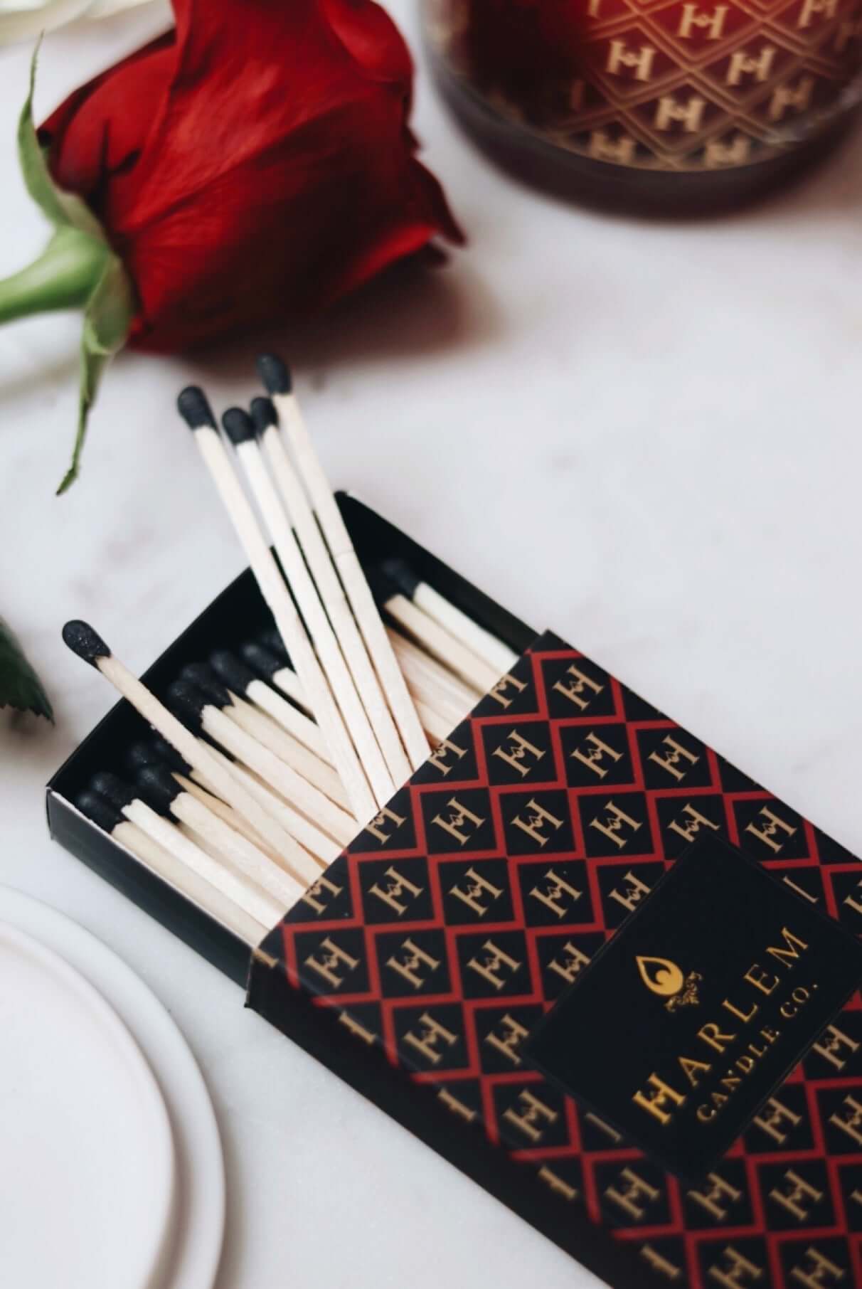 Red, black and gold Match box containing 3 inch matches with black tips.  The box has the Harlem H pattern in an art deco style printed on the box.  The image is on next to flowers.
