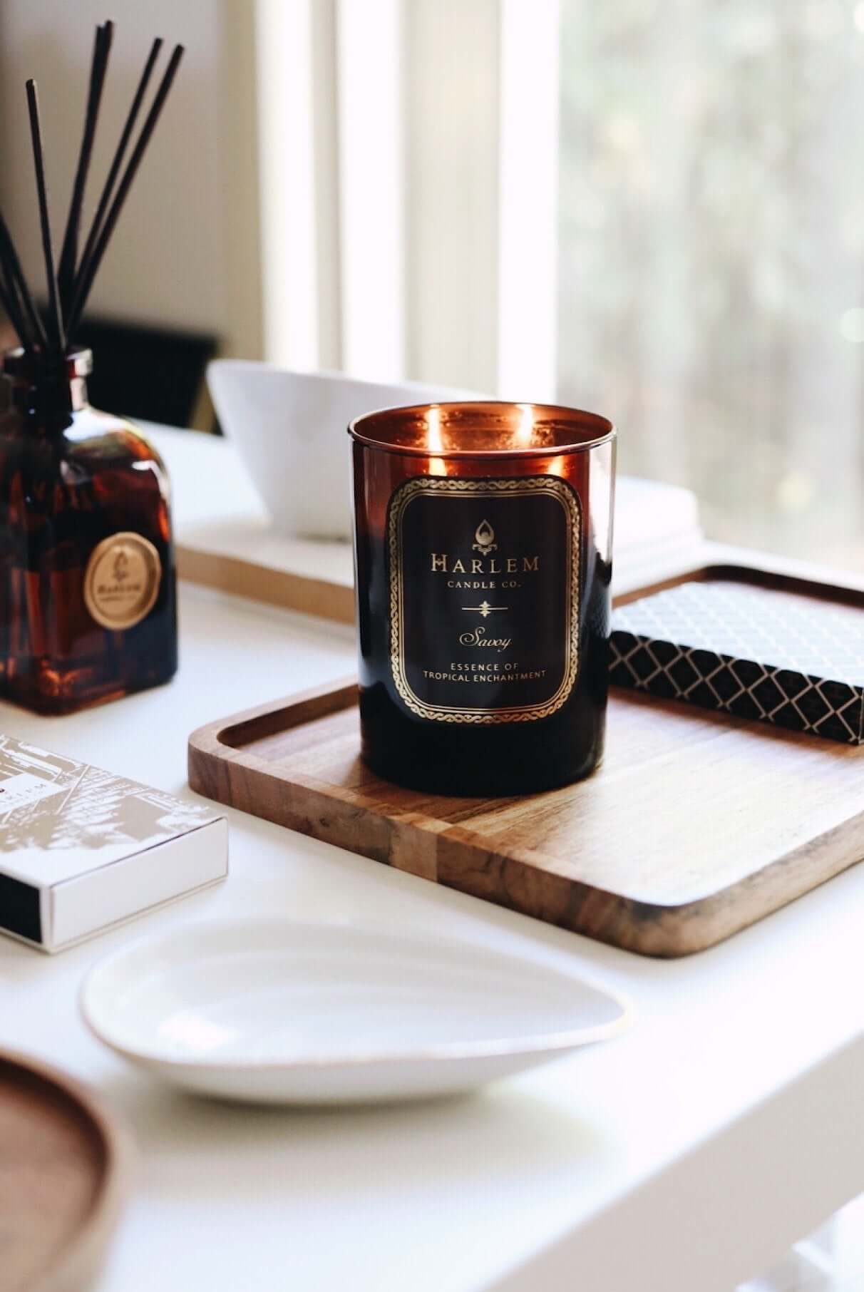 This is our Amber Savoy 12 oz 2 wick lit candle sitting on a wooden tray on a white table with our Savoy Diffuser and matches in the background.