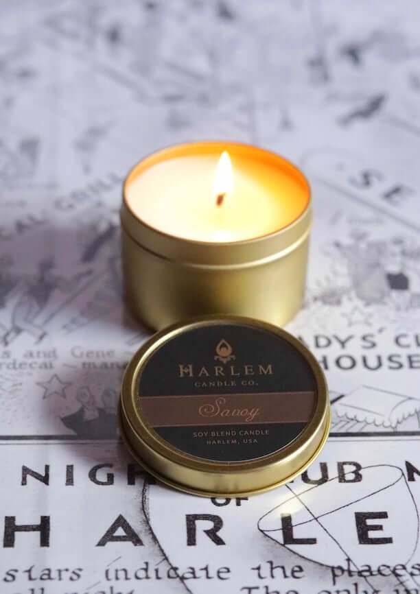 A lifestyle image of our Our stunning Savoy travel candle in a  gold metal tin. The candle has a soft flame and sits atop our black and white Harlem Map.