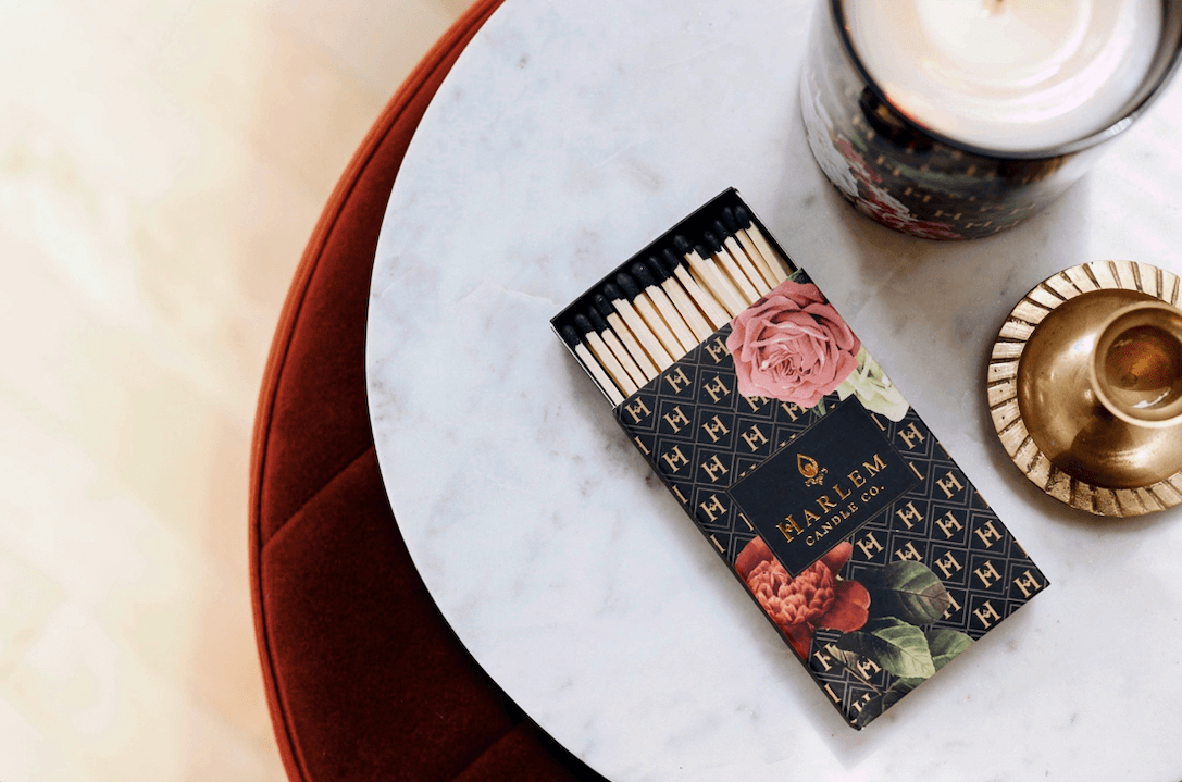 A gorgeous lifestyle image of our Black and Gold H Pattern Floral Art Deco match box with 3 inch matches with black tips opened and sitting next to a candle. 