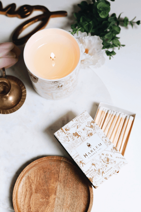 A stunning lifestyle image of Our stunning 3 inch matches with white tips, encased in our white and gold nightclub map of harlem match box on a table surrounded by leafy greens, flowers and a lit candle.